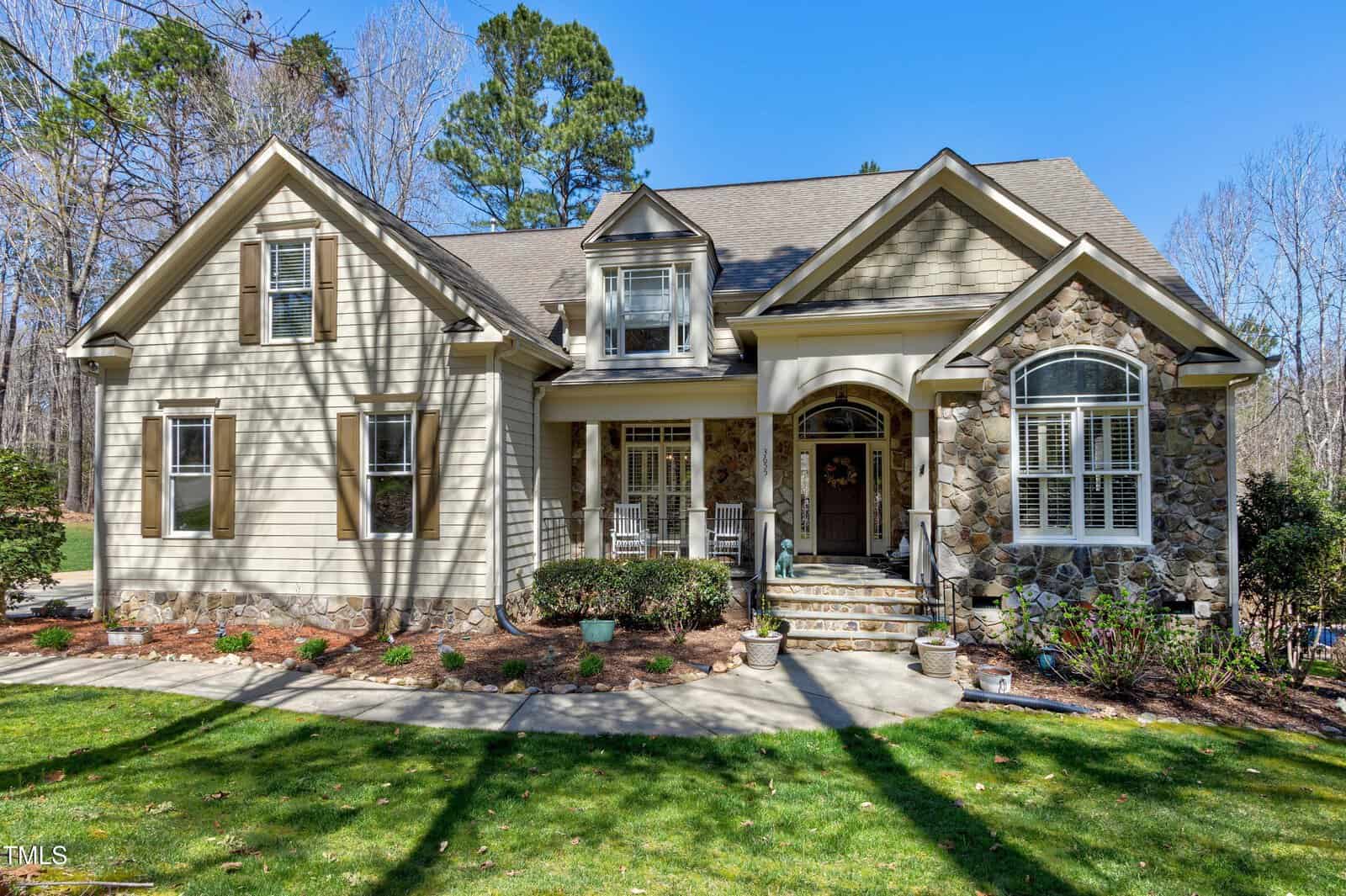 Located at 3655 Pleasants Ridge Drive in Wake Forest, this tan house with stone accents features a gable roof and white trim. A cozy front porch with columns and railings greets you, while a curved walkway winds through the landscaped yard surrounded by leafless trees.