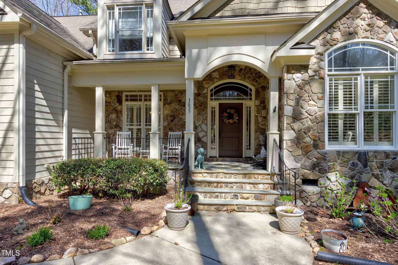 A stone and wood house with a covered porch featuring two rocking chairs and potted plants. The porch has stone steps and a wreath on the front door. There are small shrubs and decorative stonework on the facade.