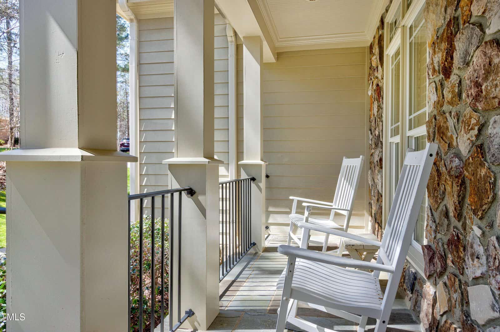 A front porch with two white rocking chairs and a small table. The porch features stone and wood elements with a black railing, and overlooks a grassy area with trees in the background.
