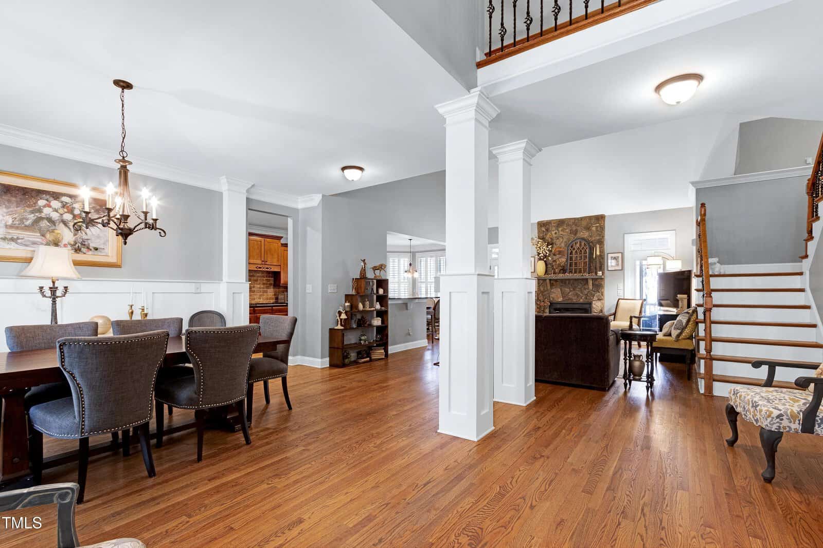 A spacious interior with hardwood floors features a dining area with a table and chairs on the left. A living room with a fireplace is to the right. Stairs with a wooden railing lead to an upper level. Walls are light-colored, and a chandelier is above the table.