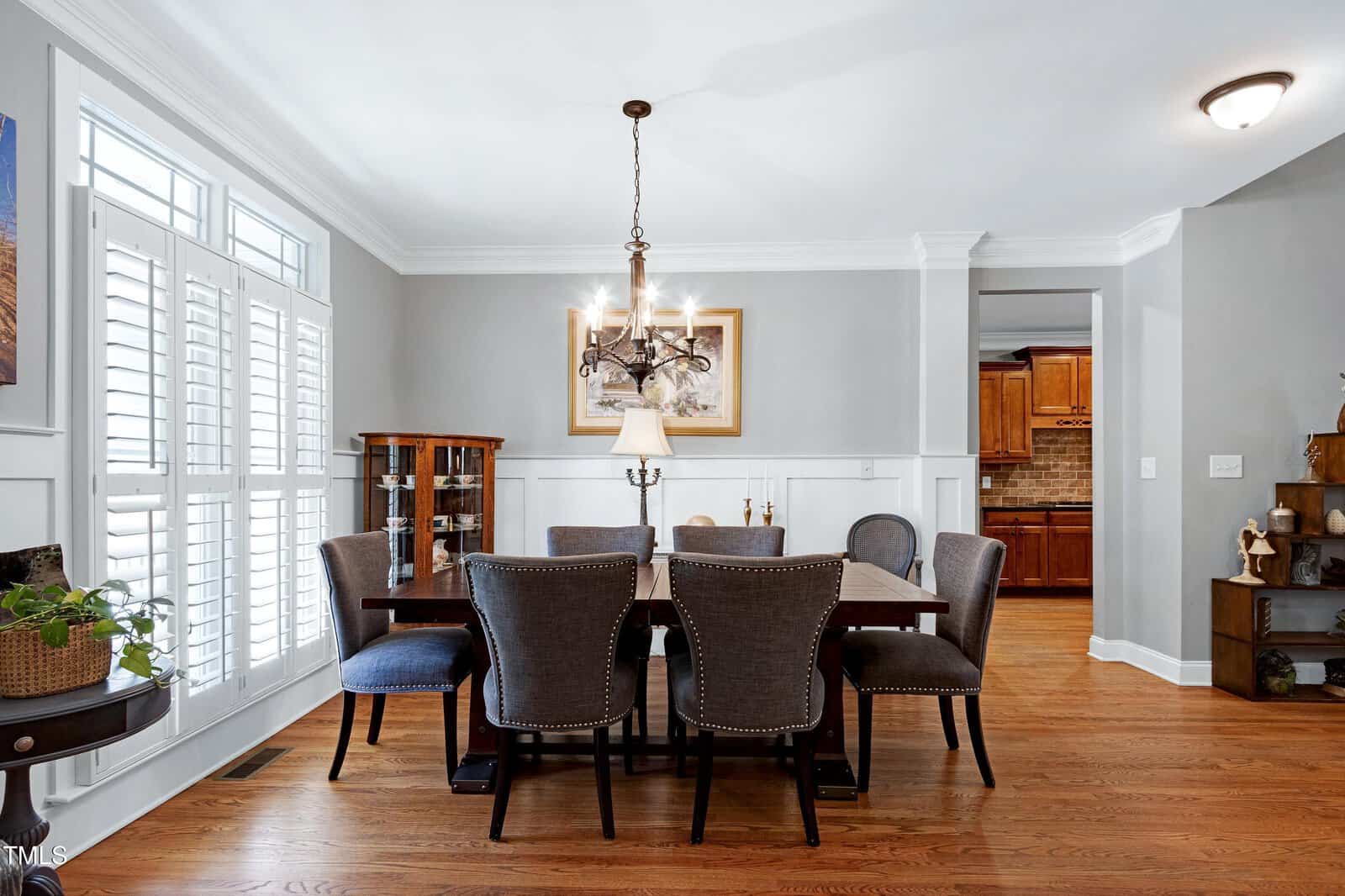 A dining room with a wooden table and eight upholstered chairs. A chandelier hangs above the table. The floor is hardwood, and there is a cabinet in the corner. Large windows with shutters are on the left, and the adjacent room features wooden cabinets.