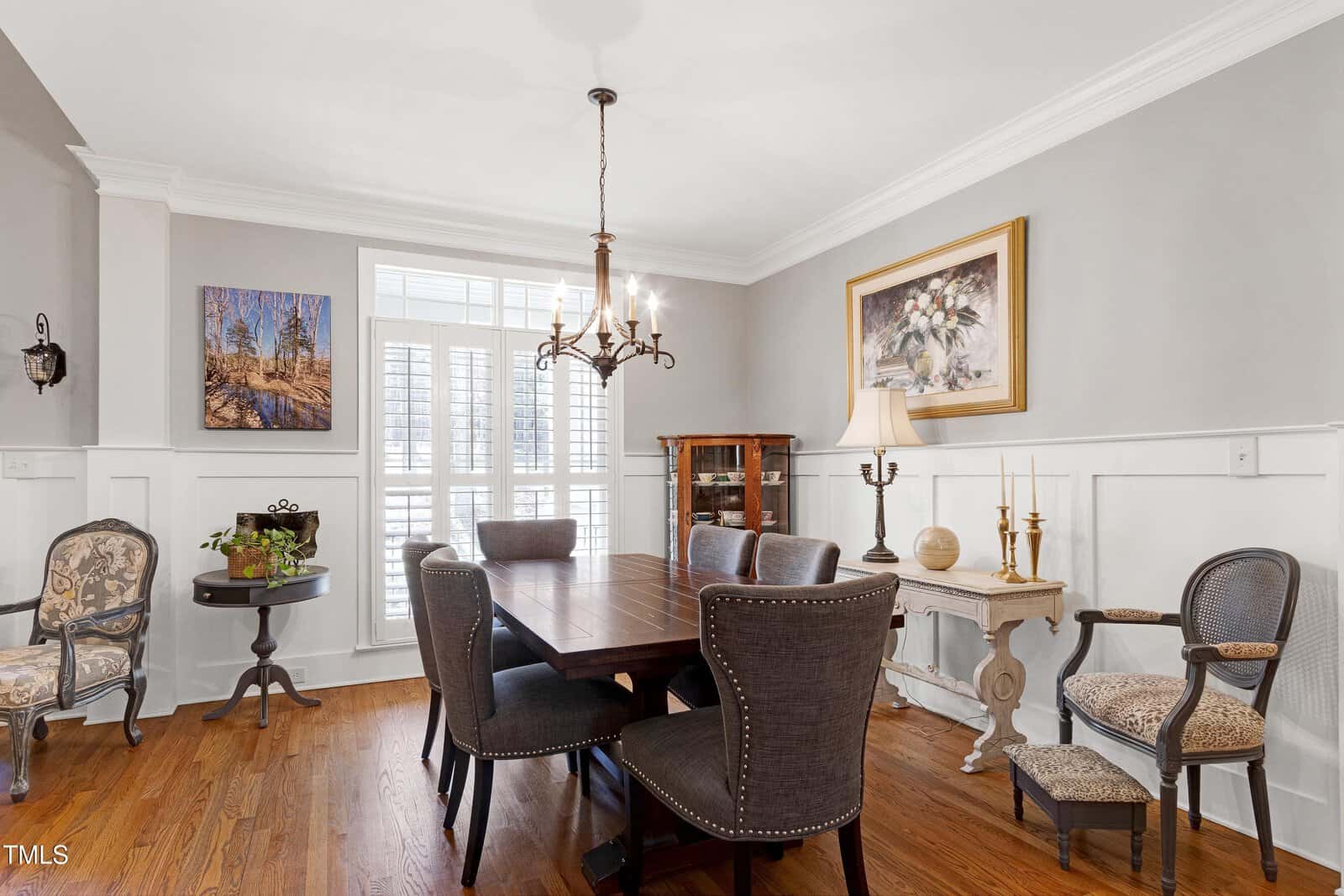 A dining room features a dark wooden table with six upholstered chairs. A chandelier hangs above the table. The walls are light gray with white paneling. There are decorative chairs, framed pictures, and a side table with a lamp and candles.