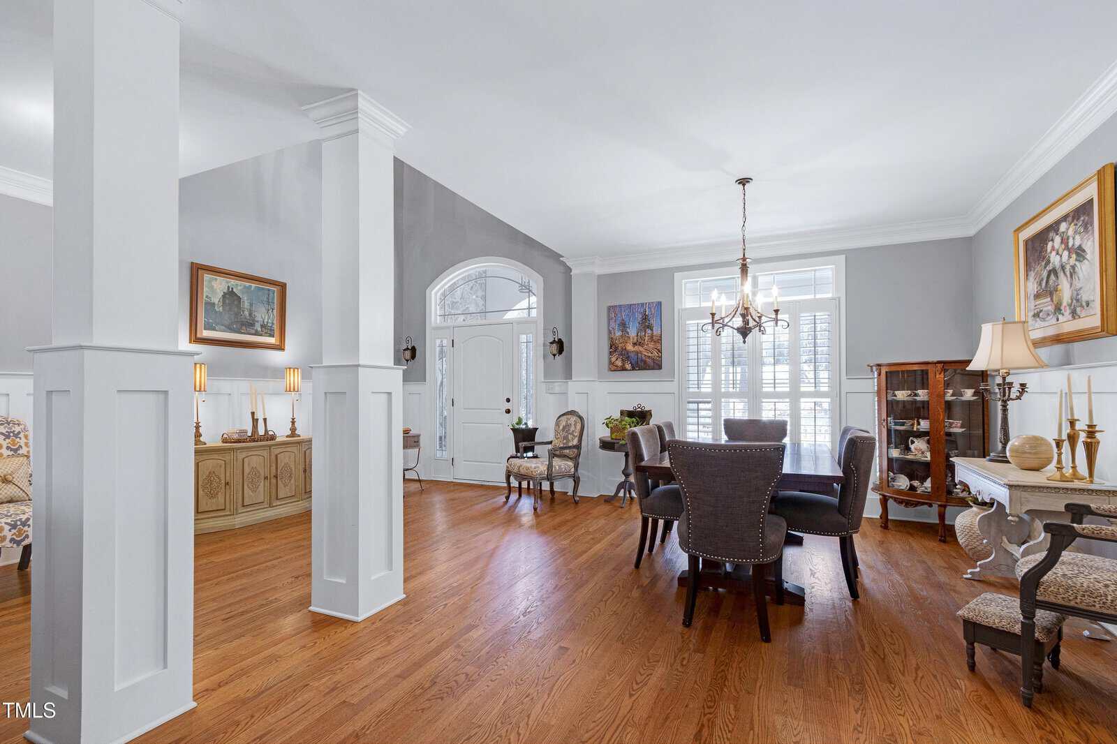A dining room with a round table and six chairs on wooden flooring. The room has light grey walls, a large white-framed window, and a chandelier. Decorative cabinets, paintings, and a seated statue add to the decor. An archway leads to another room.