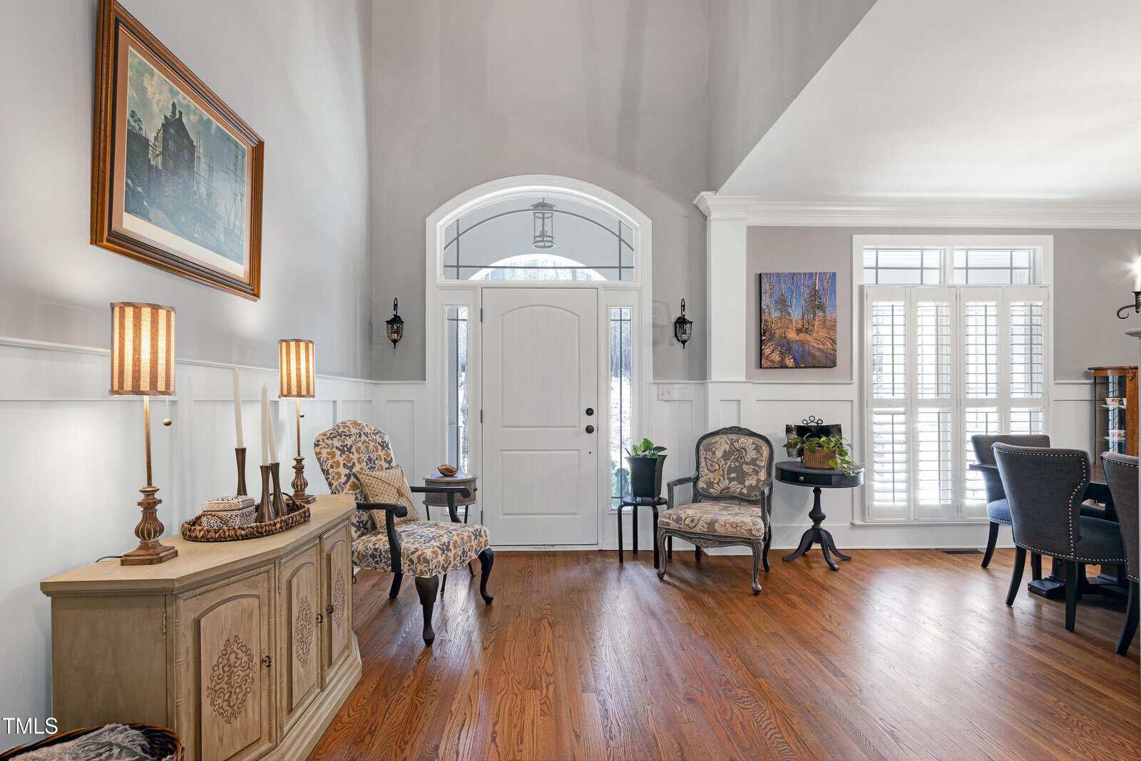 A bright living room with a wooden floor features a white front door with an arched window above. Two patterned chairs and small tables are by the door. Wall art decorates the space, which is lit by natural light through large windows with shutters.