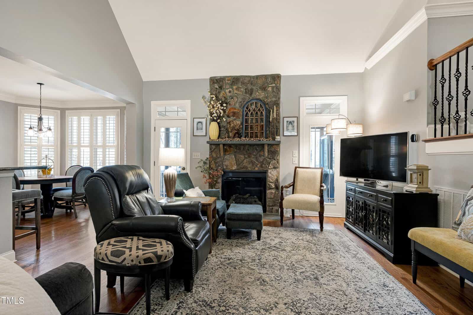 A cozy living room with a high ceiling features a stone fireplace with a decorative mirror and vases on the mantel. There's a black leather armchair, patterned rug, TV, and a dining area in the background. Natural light filters through wide windows.