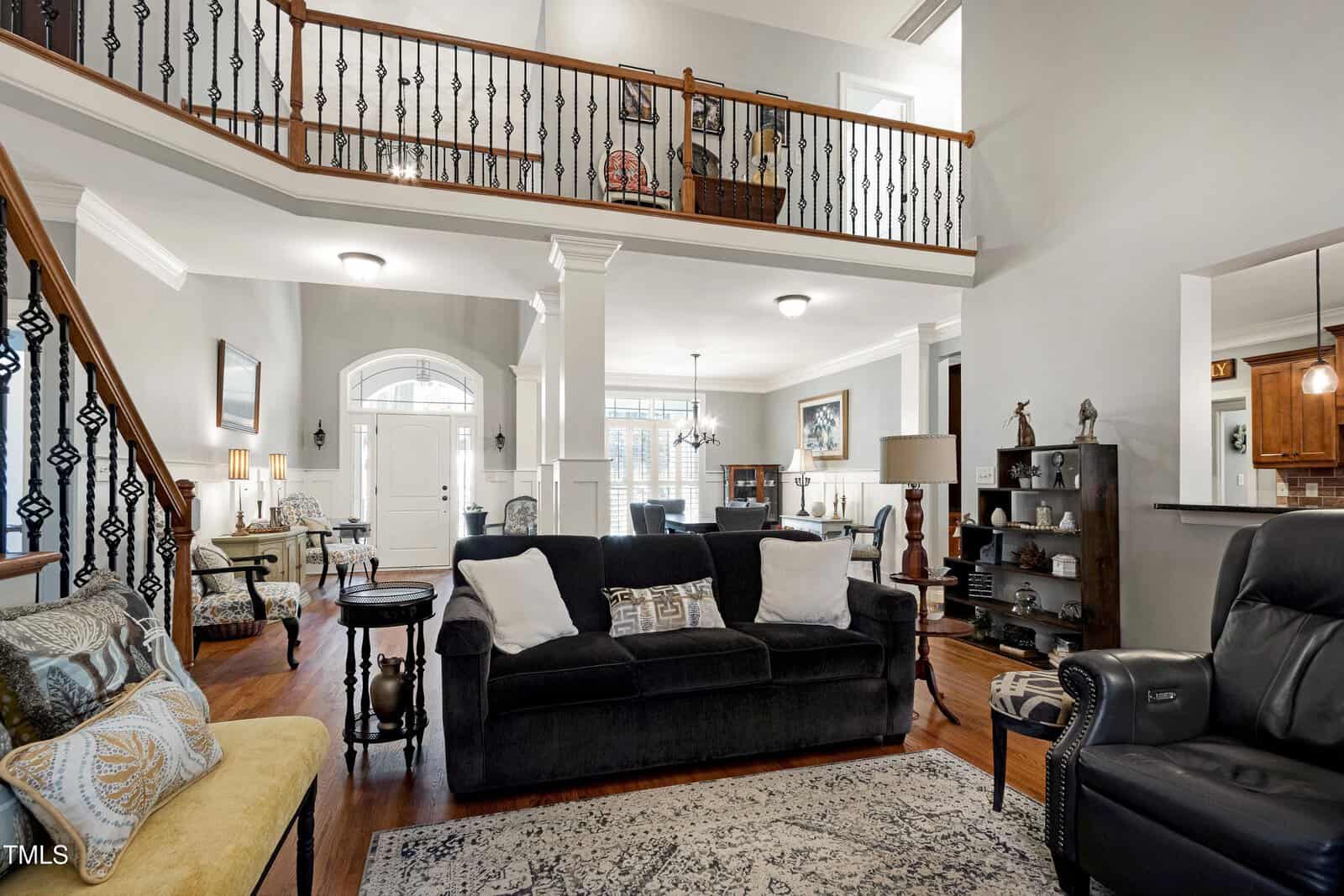 A spacious living room with high ceilings and a mezzanine. It features a black sofa, a cushioned bench, armchairs, and a black recliner. The room has hardwood floors, a patterned rug, and various decorative items. A dining area is visible in the background.