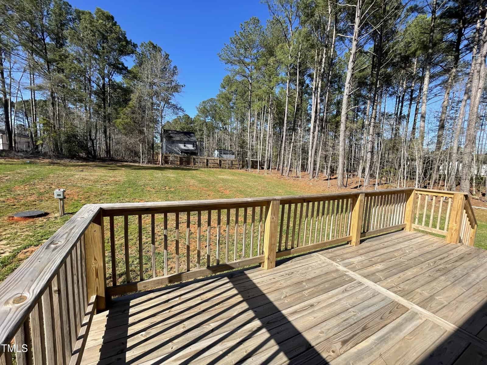 A wooden deck overlooks a spacious backyard with patchy grass and tall pine trees. A house is visible in the background, and the sky is clear and blue.