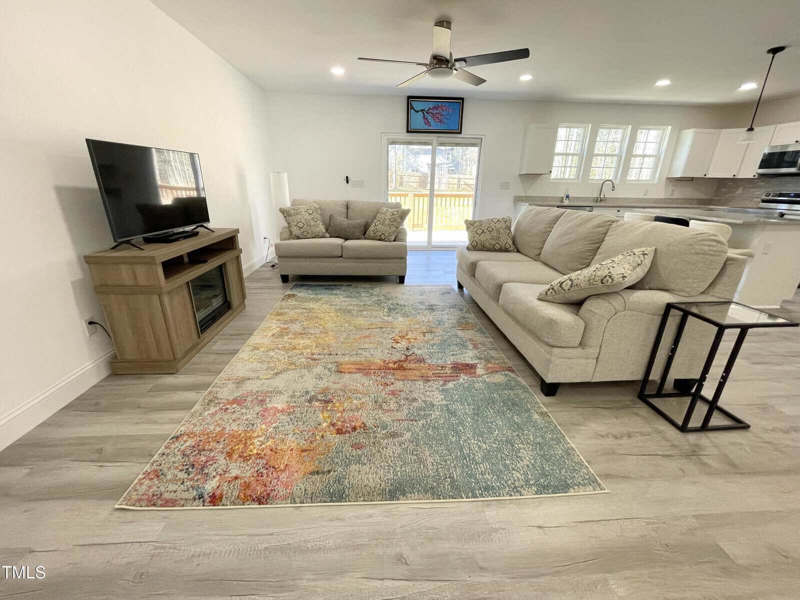 A living room with two beige sofas, a patterned rug, a wooden TV stand with a TV, and a side table. The room has light wood floors, white walls, ceiling fan, and an open kitchen in the background.