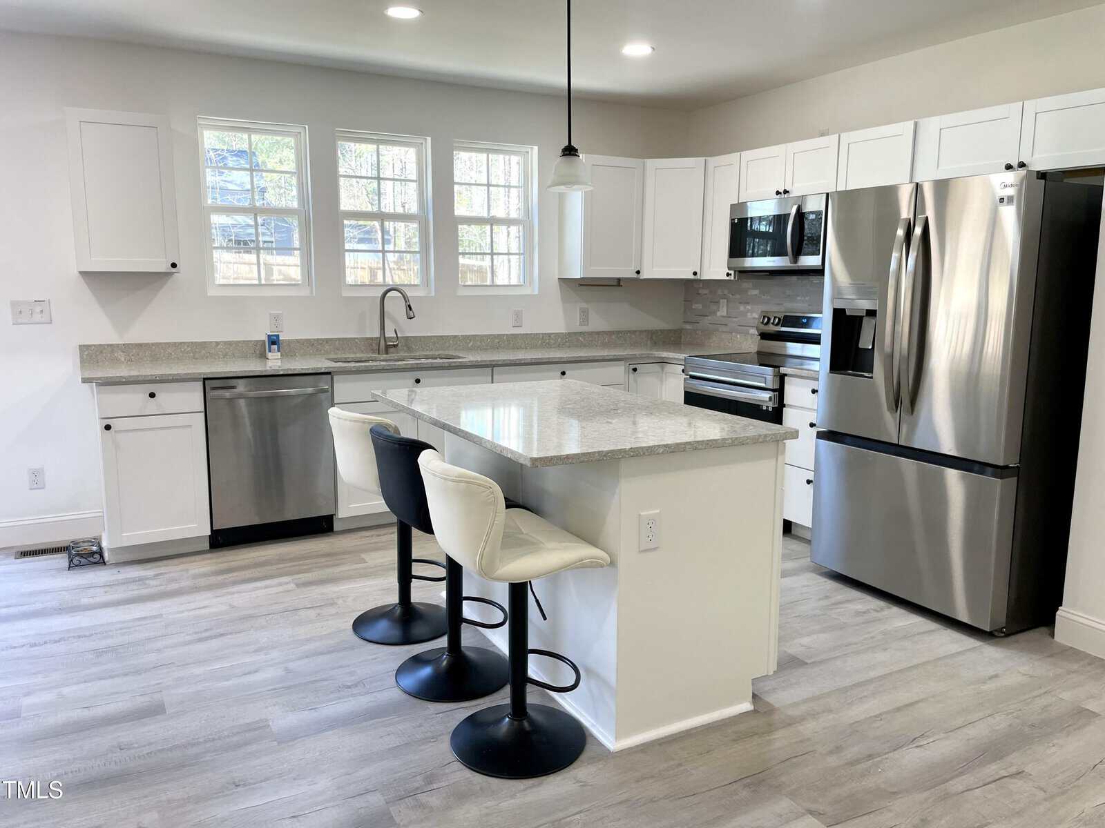 Modern kitchen with white cabinets, stainless steel appliances, a center island with two cream barstools, light gray countertops, and wood-look flooring. Three windows above the sink let in natural light.
