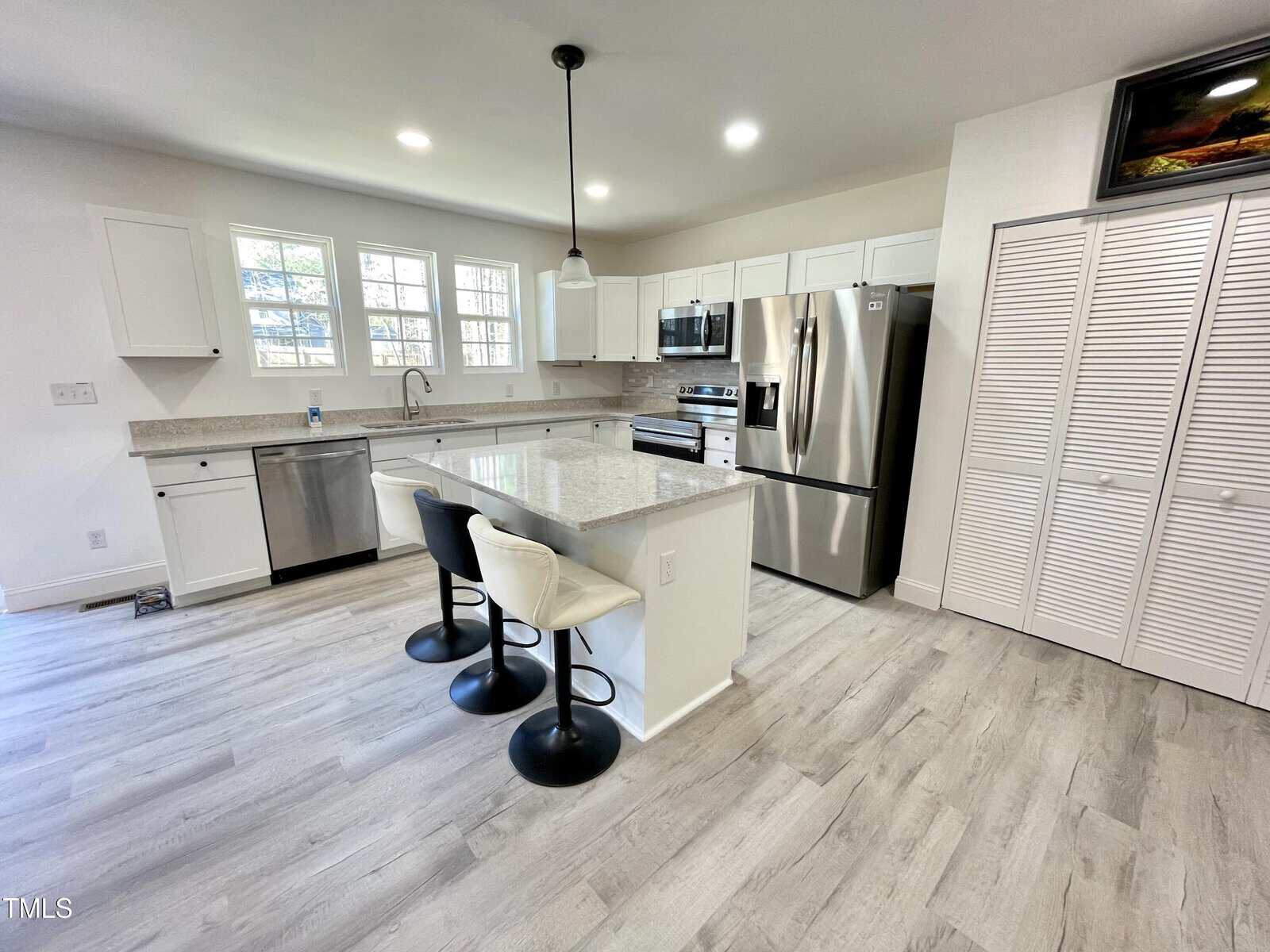 Modern kitchen with white cabinets, stainless steel appliances, a center island with three barstools, pendant lighting, light wood flooring, and large windows letting in natural light.