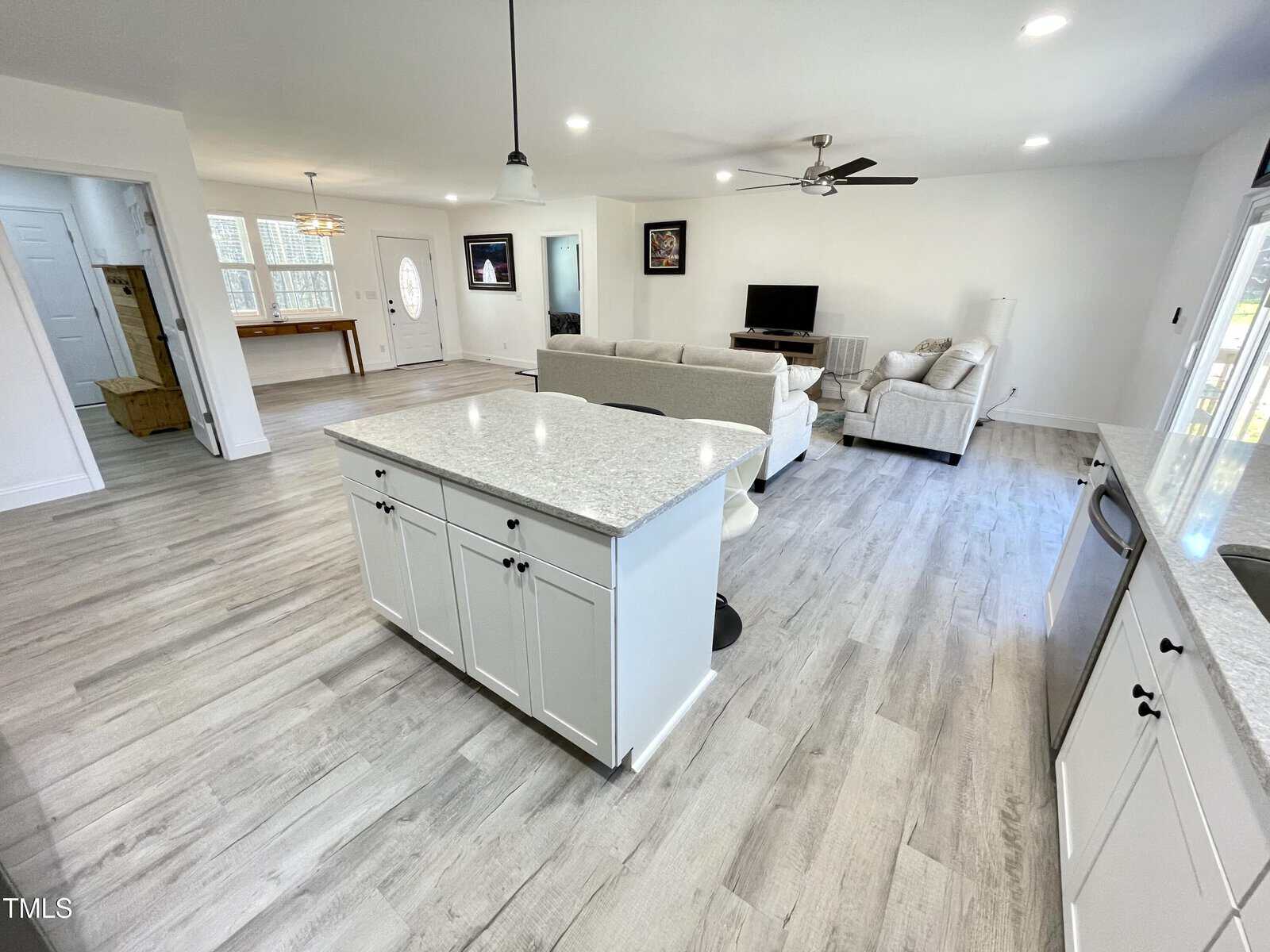 Open-concept living area with light wood floors, white cabinets, and a kitchen island in the foreground. A living room with two couches, TV, and ceiling fan is visible, along with an adjacent dining area and entryway.