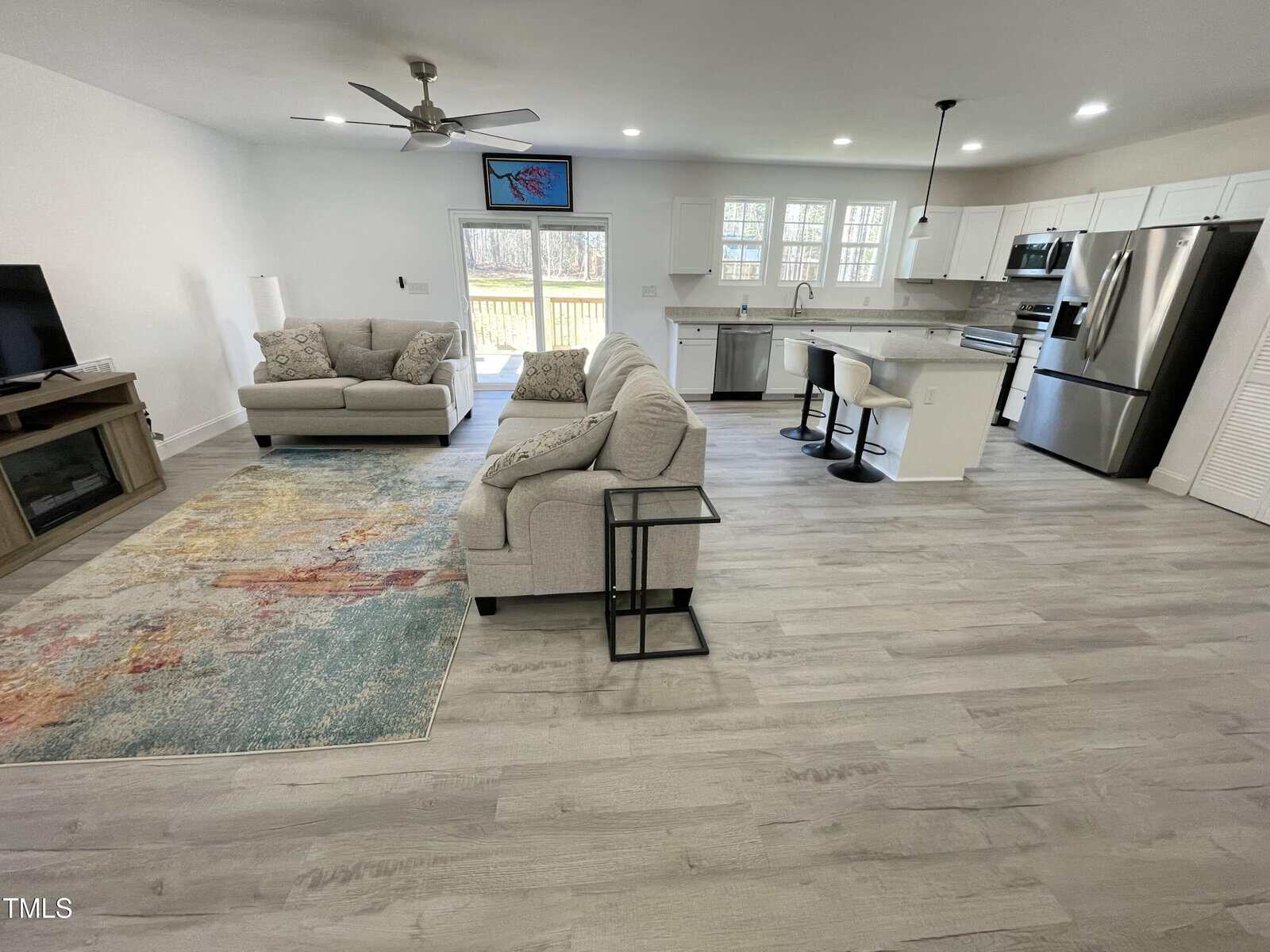 Open-concept living room and kitchen with light gray wood flooring, two light gray sofas, a colorful area rug, flat-screen TV, white cabinetry, stainless steel appliances, and a breakfast bar with two stools.