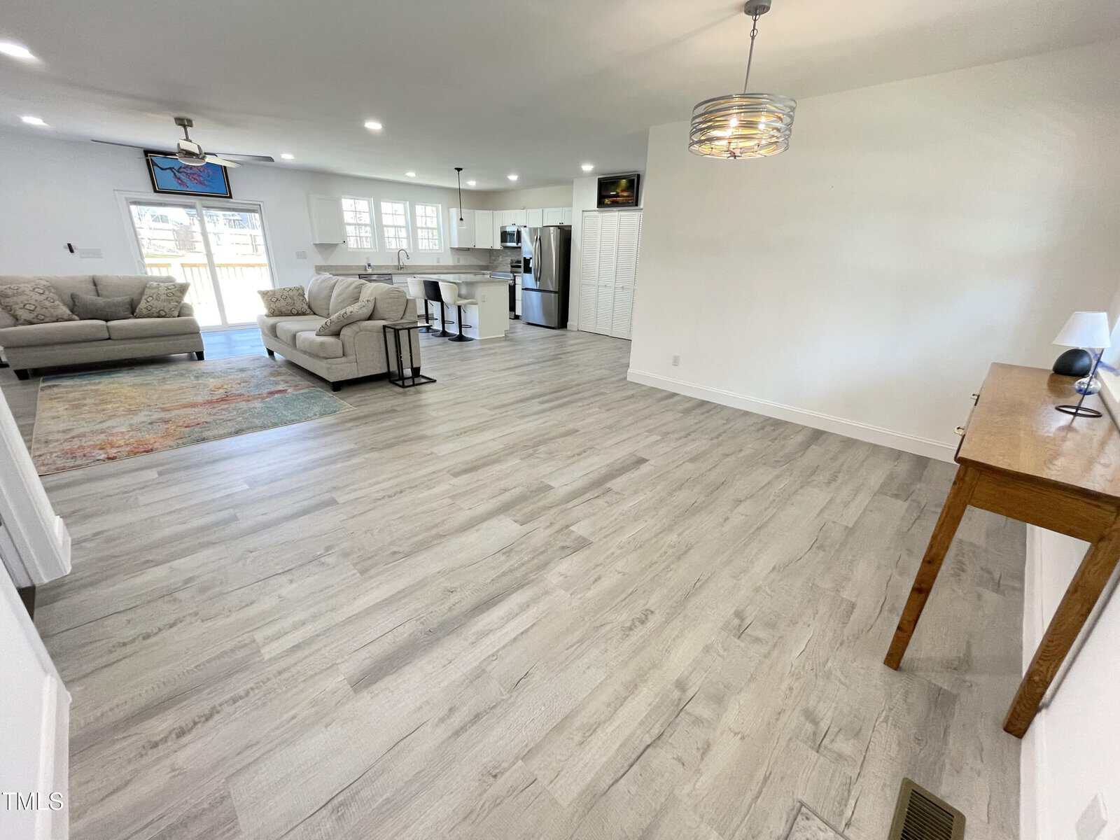 Spacious open-concept living area with light wood flooring, two beige couches, a colorful rug, and a kitchen in the background. A wooden console table with a lamp sits against the right wall. Large windows provide natural light.