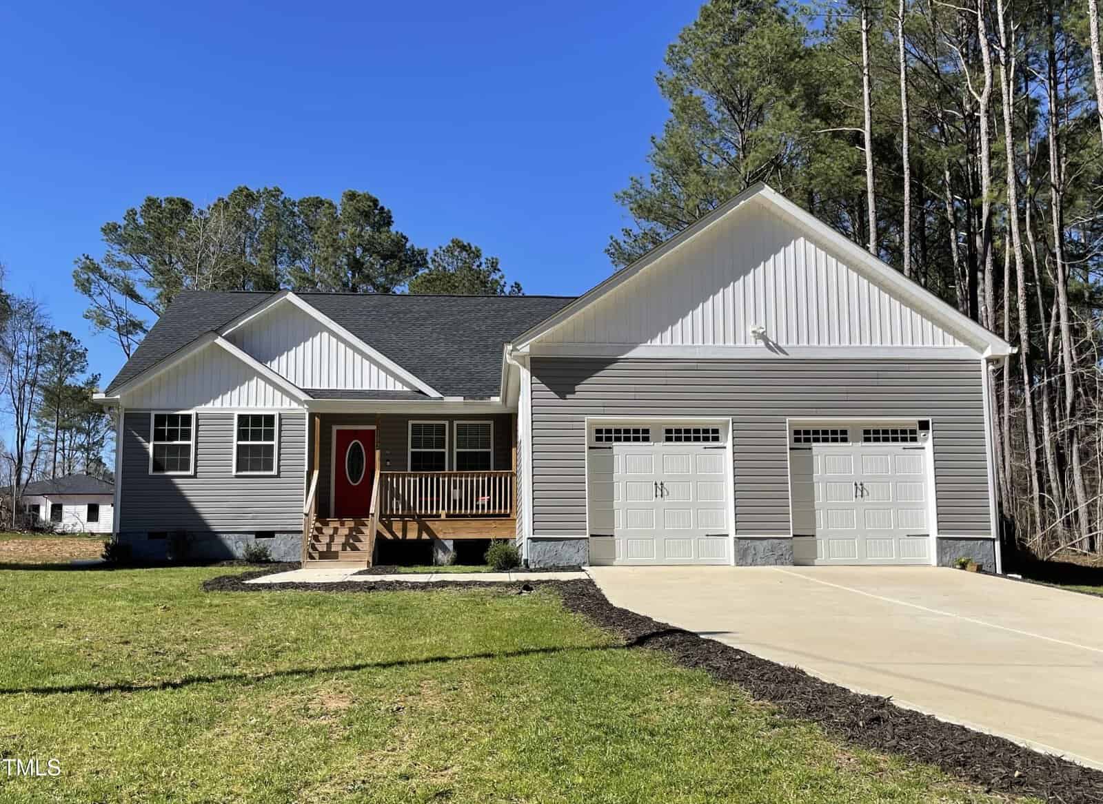 Single-story house with gray siding, a red front door, a small porch with wooden steps, and a two-car garage. Concrete driveway and green lawn in front; tall trees are in the background under a clear blue sky.