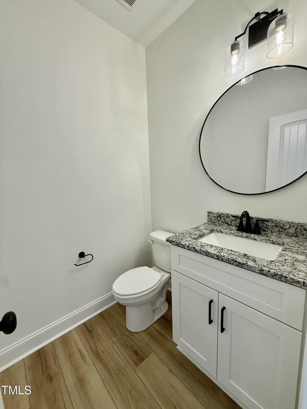A small bathroom with wood-look flooring, a white toilet, a white vanity with a granite countertop, a black faucet, a round mirror, and a wall-mounted light fixture above the mirror.