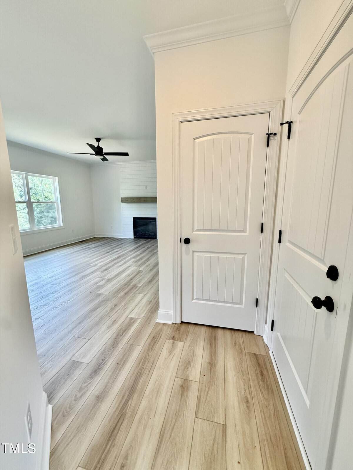 A hallway with light wood flooring leads to a living room with large windows, a ceiling fan, and a fireplace. The hallway features white paneled doors with black handles and white walls.