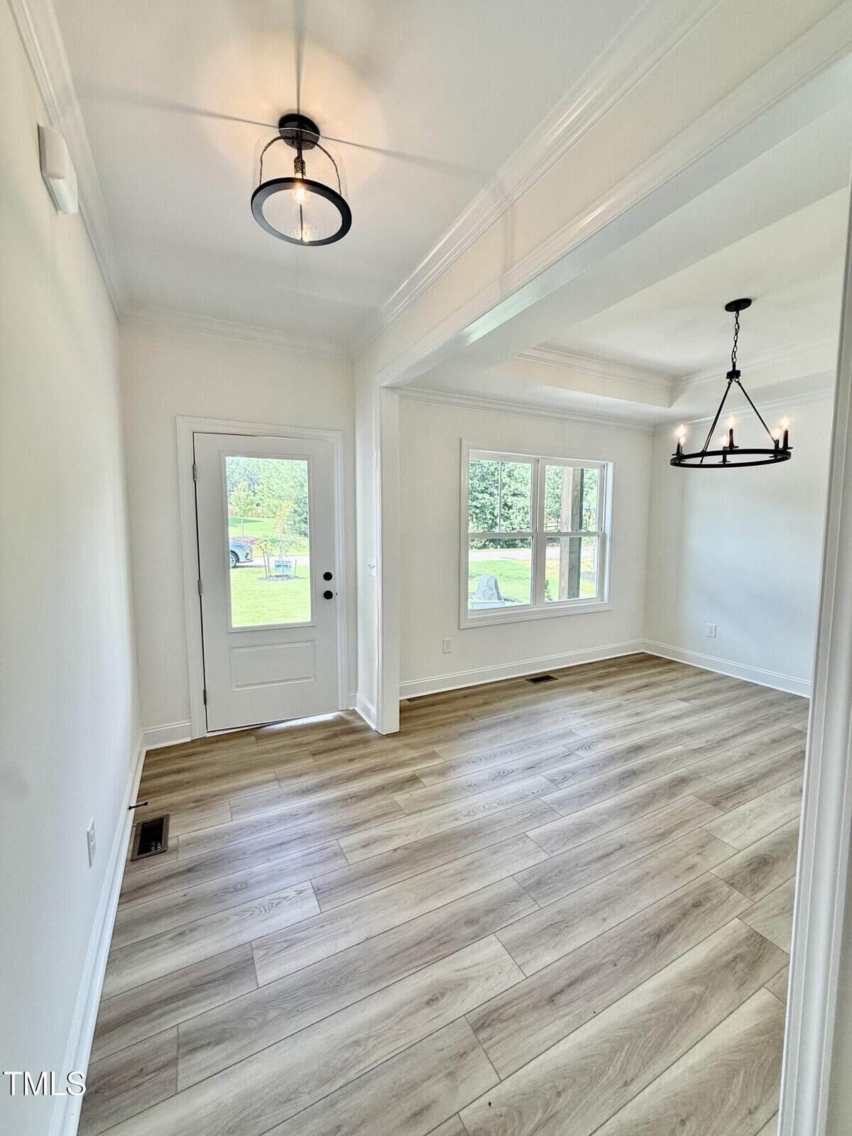 A bright room with light wood flooring, white walls, a front door with glass panels, and two modern black light fixtures. Three windows provide natural light and a view of a green yard outside.