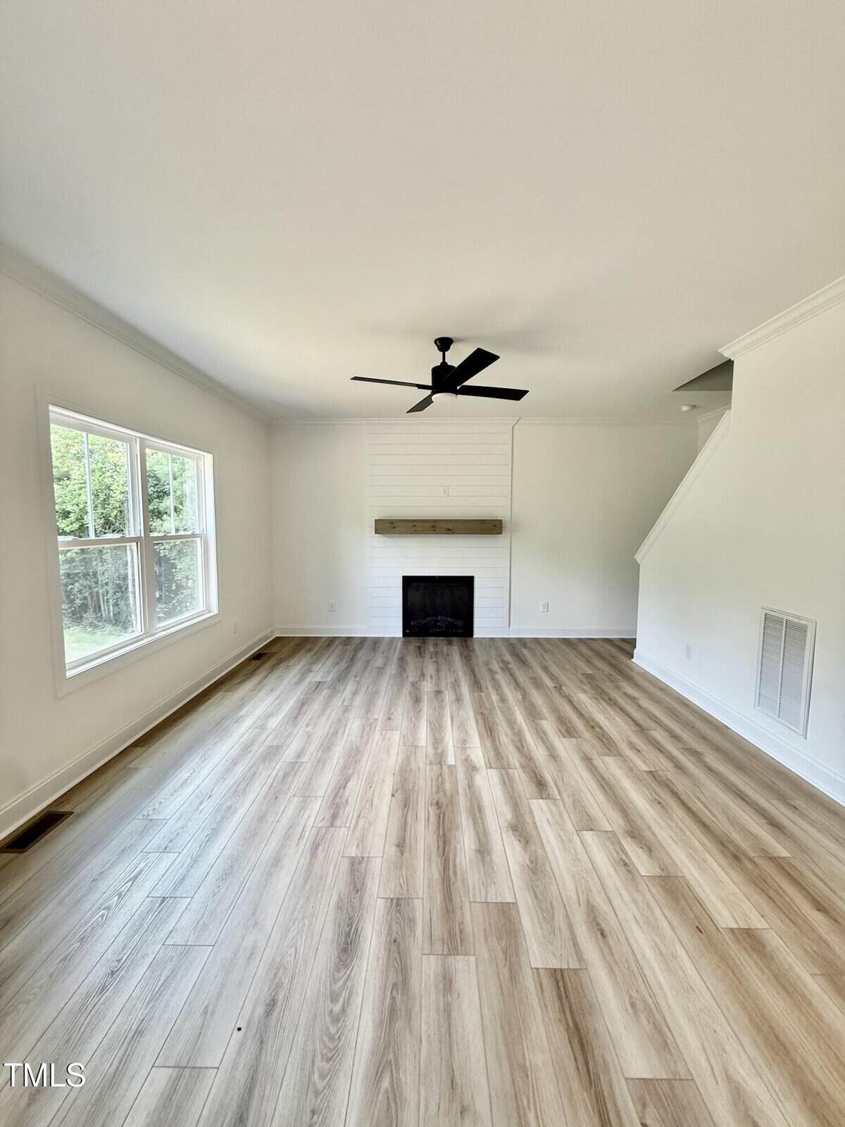 A bright, empty living room with light wood flooring, large windows on the left, a black ceiling fan, a fireplace with a wooden mantel, and stairs ascending on the right side. Walls are painted white.