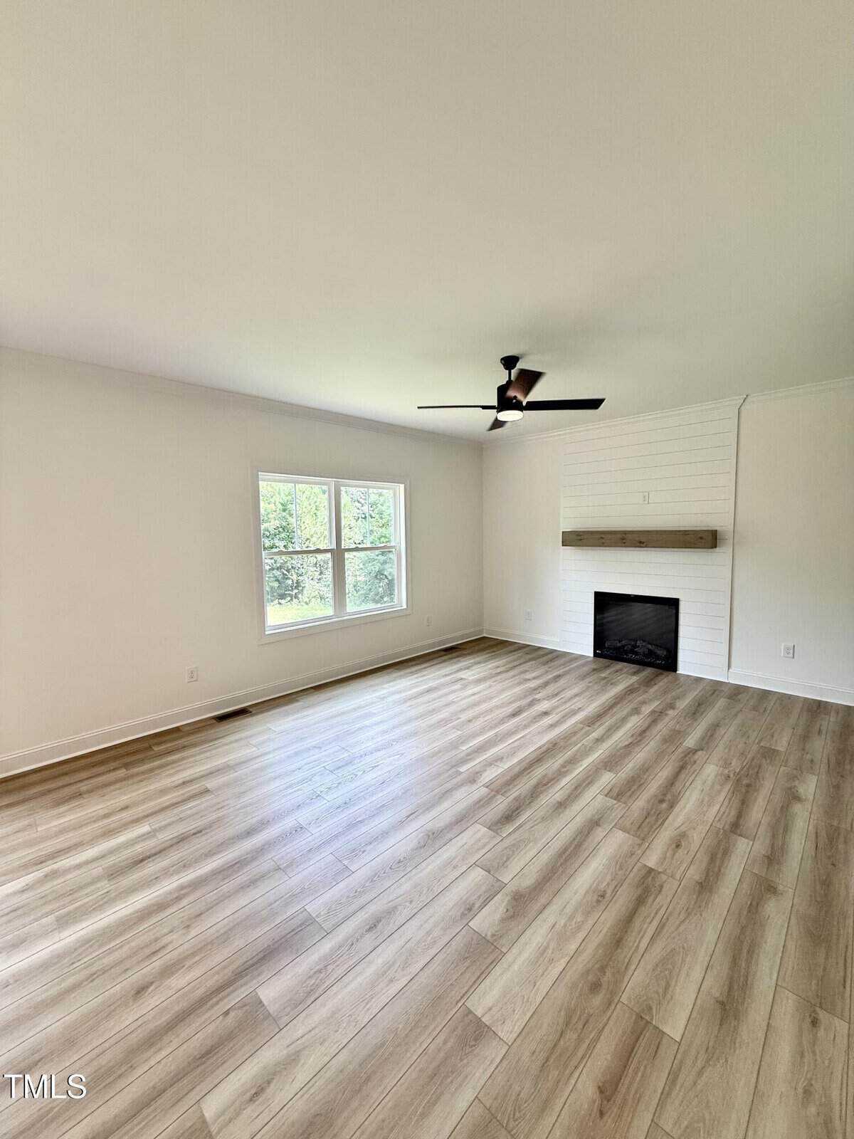 A bright, empty living room with light wood flooring, a ceiling fan, a window on the left wall, and a white shiplap accent wall with a built-in fireplace and wooden mantel.