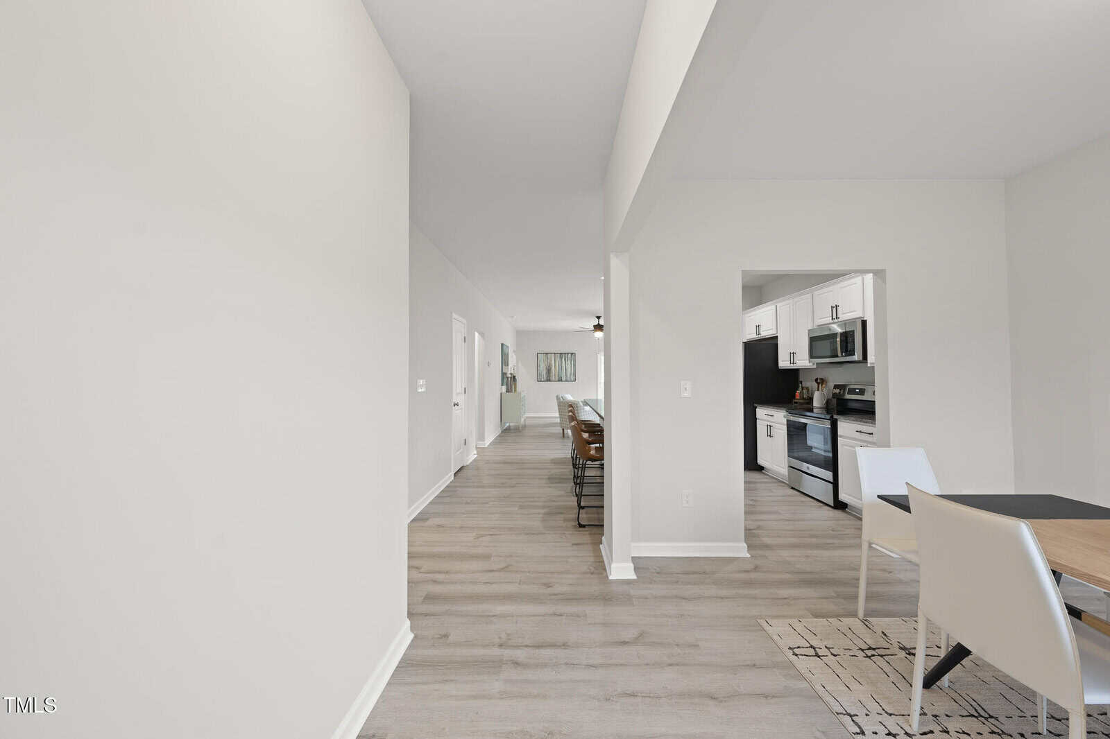 A modern, open-concept home interior with light-colored walls and wood flooring, a dining table with white chairs, a kitchen with stainless steel appliances, and a hallway leading to other rooms.