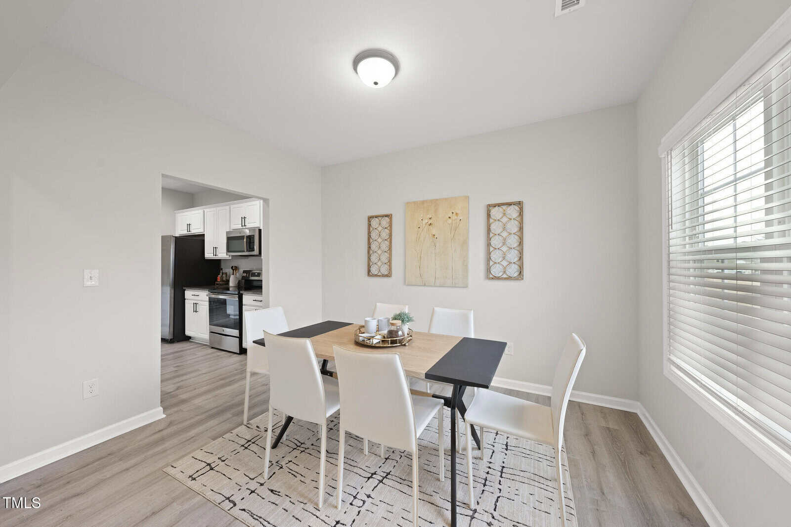 A dining area with a wooden table, six white chairs, and a patterned rug. The walls are light-colored, and there are three framed artworks. A window with blinds lets in natural light, and an adjacent doorway leads to a kitchen.