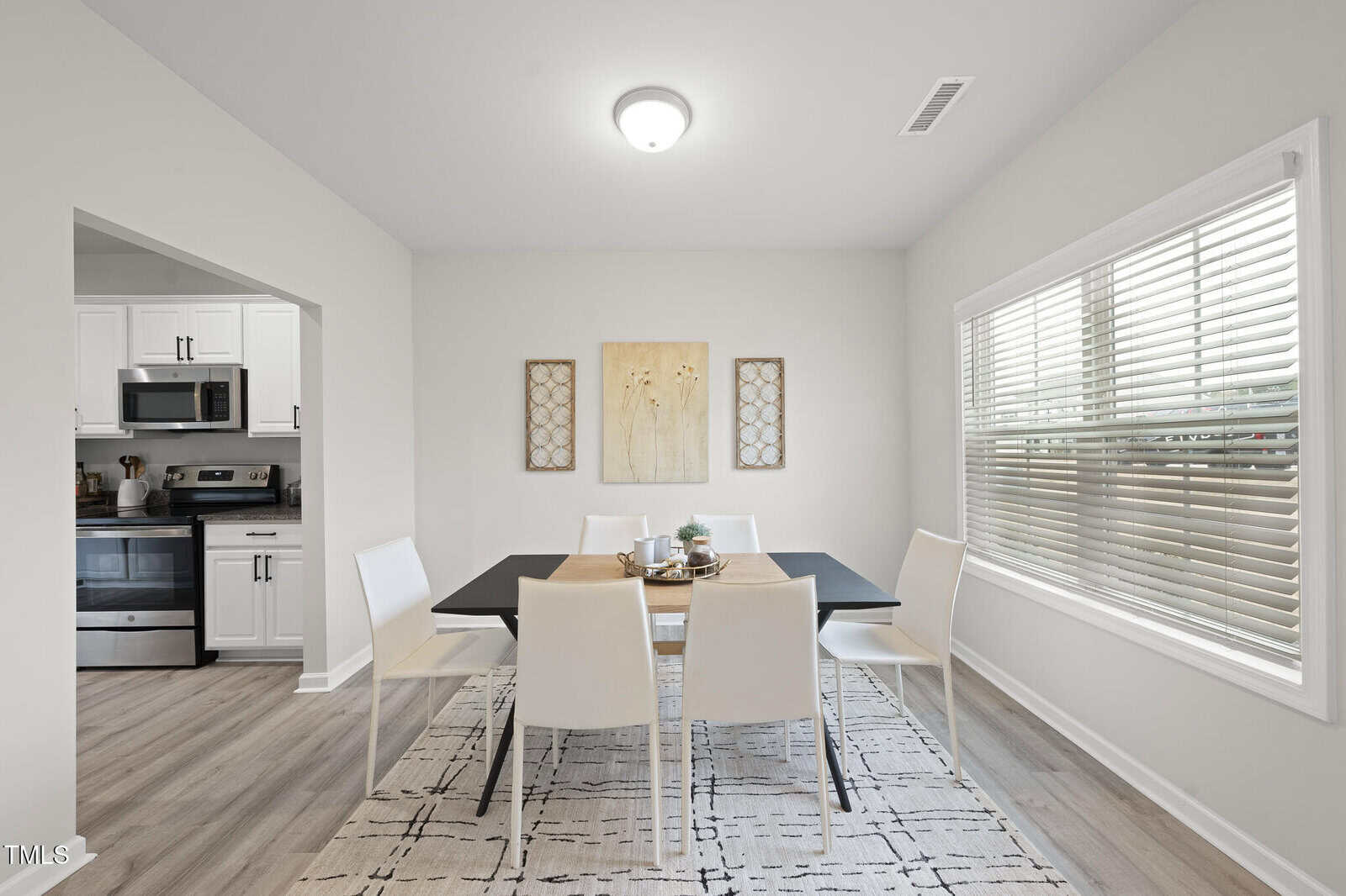 A dining area with a black rectangular table, six white chairs, a white and black rug, wall art, and a large window with blinds. An adjoining kitchen with white cabinets and a stainless steel stove is visible.