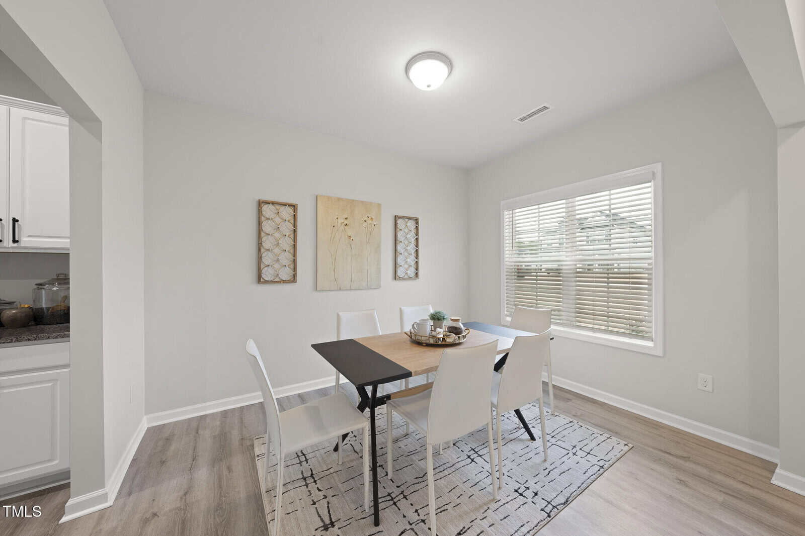 A modern dining room with a rectangular table surrounded by six white chairs on a patterned rug, neutral-toned walls, three wall art pieces, and a large window with blinds letting in natural light.