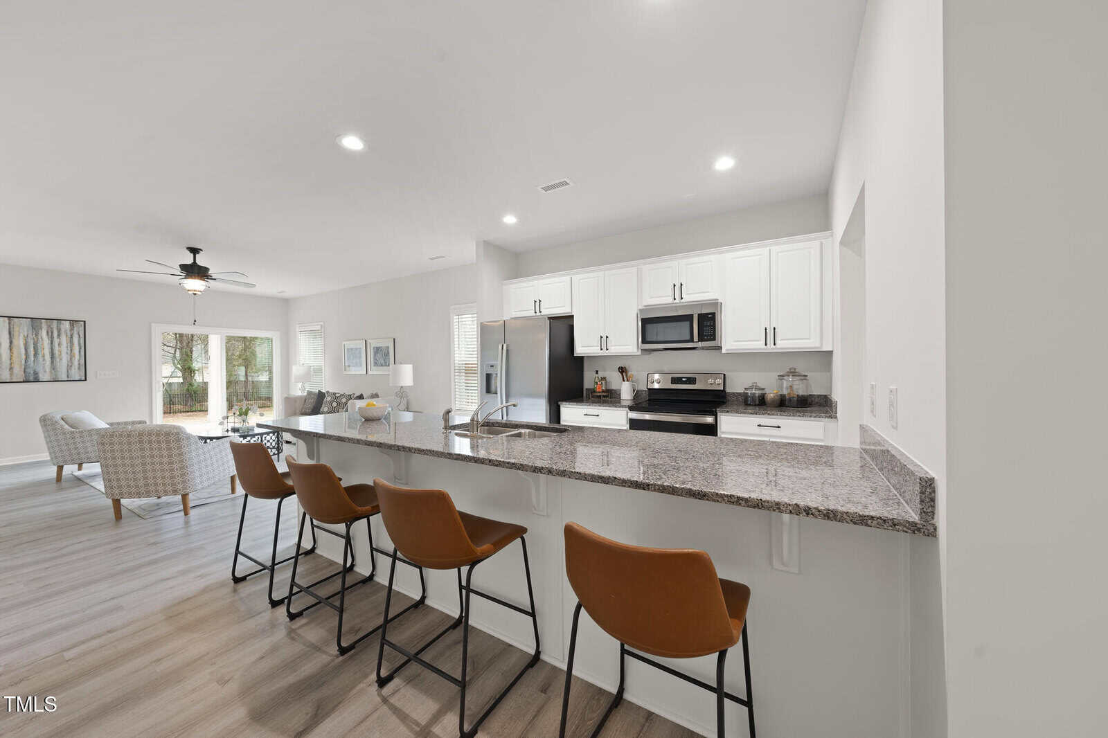 Modern kitchen with granite countertop, four brown barstools, white cabinets, stainless steel appliances, and an open layout leading to a bright living area with light wood flooring.