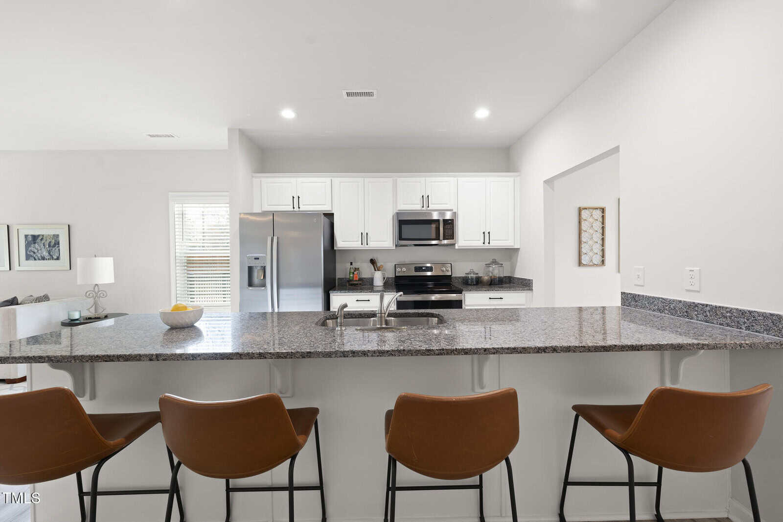 A modern kitchen with white cabinets, stainless steel appliances, a granite countertop with a sink, and four brown barstools. Decorative items and kitchen essentials are visible on the counters and shelves.