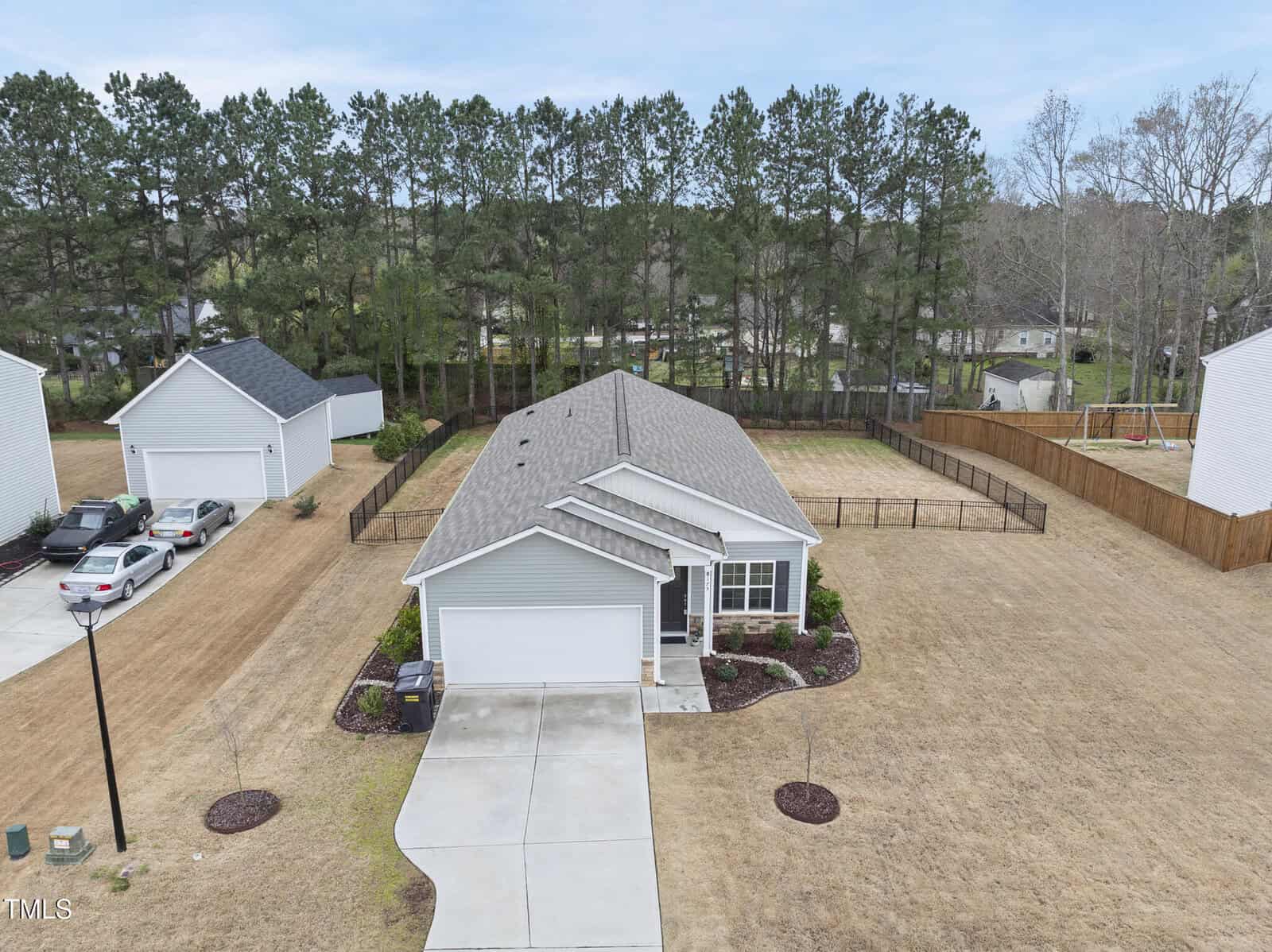 A single-story house with a gray roof and attached garage is centered in a large yard with brown grass. The property is bordered by a wooden fence, and trees line the background behind the house.