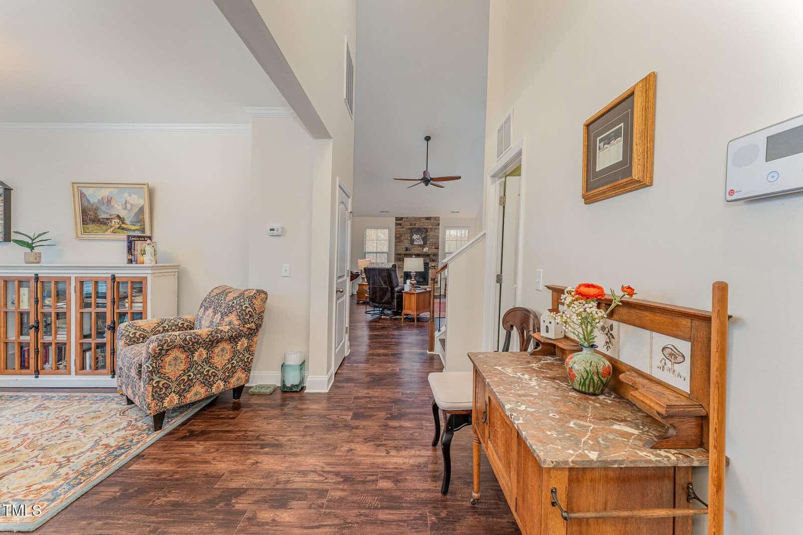 A hallway with wood flooring leads to a living area. On the right, a wooden sideboard with a marble top holds flowers and decor. A patterned armchair and a bookcase are on the left. Walls are cream-colored.