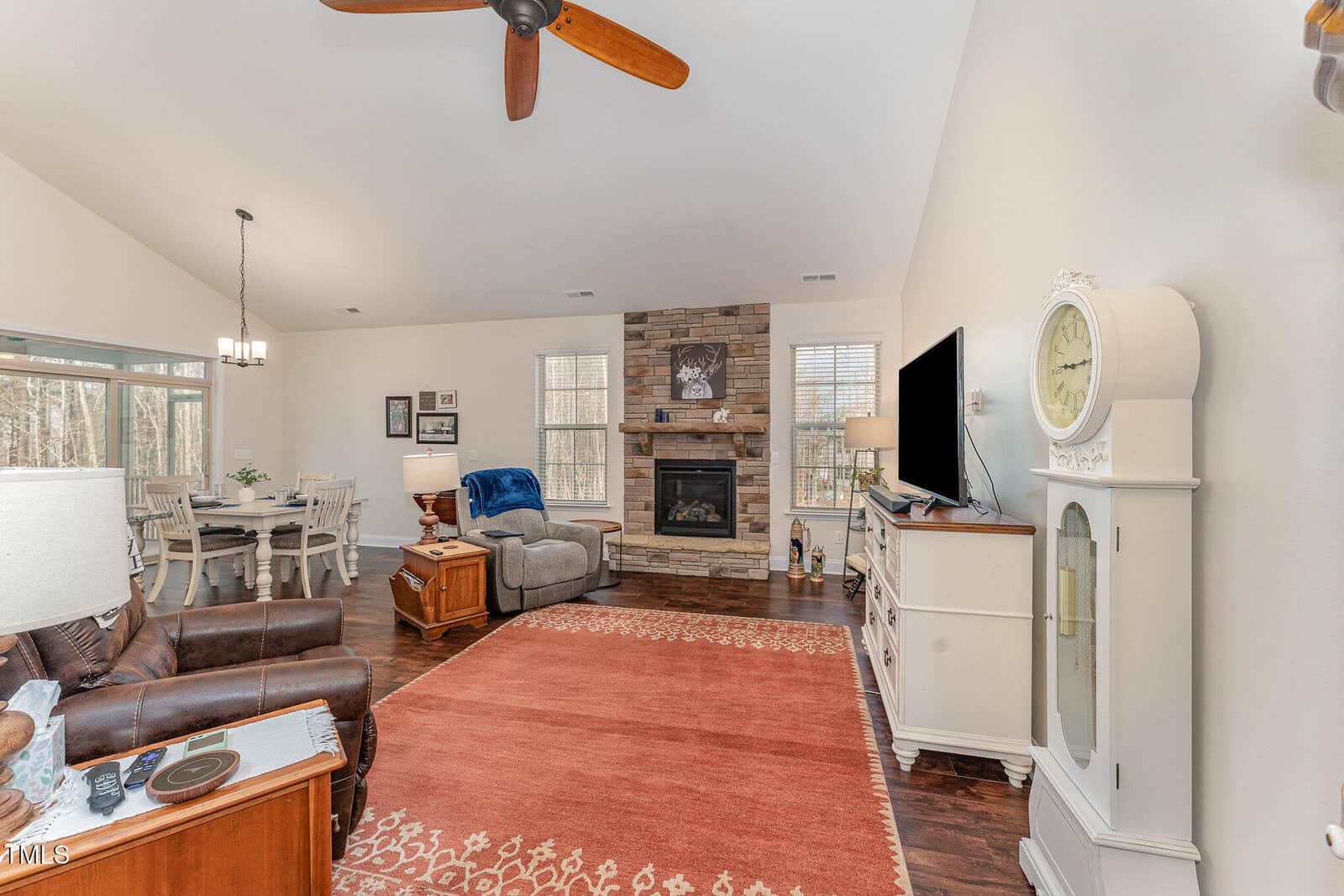 A living room with a brown leather chair, white grandfather clock, TV on a white console, stone fireplace, and a dining area with a table and six chairs, all set on wooden floors with an orange area rug.