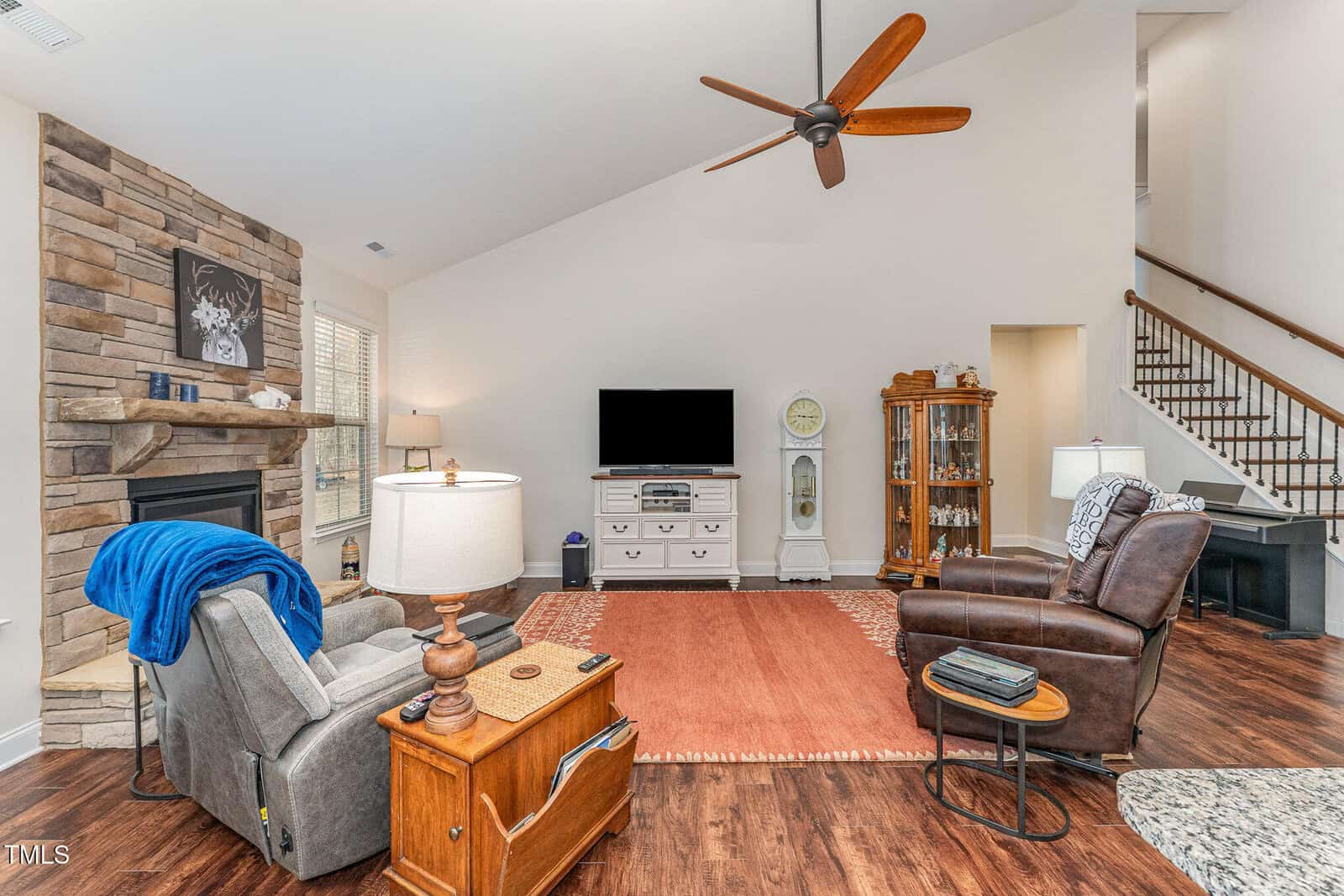 A living room with two recliners, a wooden side table, a TV on a white console, a fireplace with a stone surround, a grandfather clock, a display cabinet, and a staircase with a wooden railing.