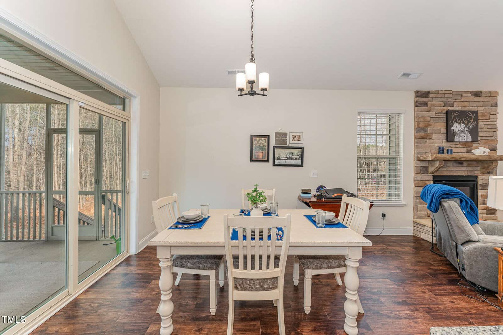 A dining area with a white table set for four, surrounded by white chairs on a wooden floor. A chandelier hangs above, with framed art on the wall, and a stone fireplace with decor is visible in the adjacent living room.