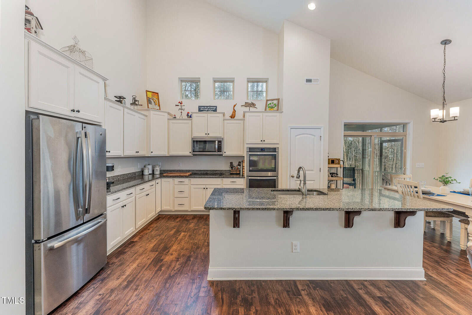 Modern kitchen with white cabinets, stainless steel appliances, a large granite island with sink, wooden floors, and high ceilings. A dining area with a table and chairs is visible to the right near large windows.