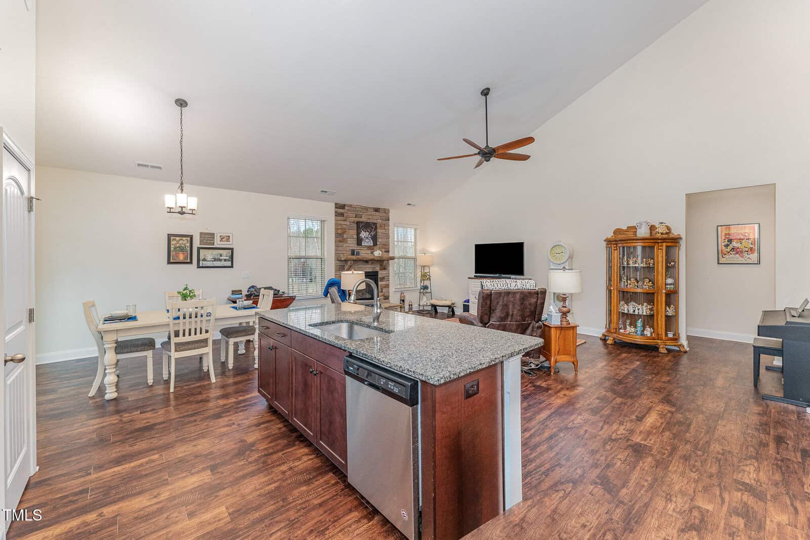 Open-concept living space with a kitchen island in the foreground, stainless steel appliances, dining table to the left, living area with armchairs, fireplace, and TV in the background, and hardwood floors throughout.