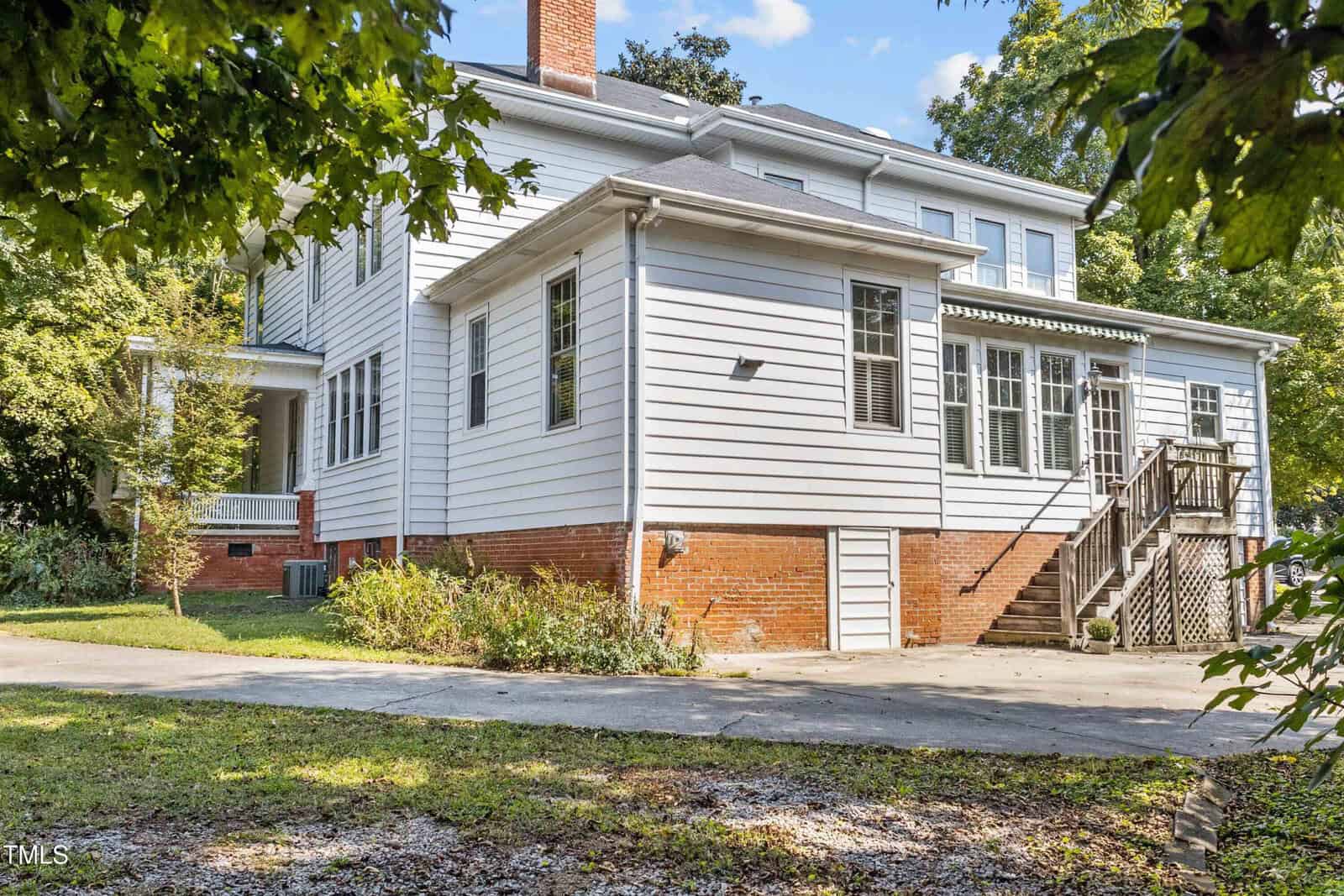 White two-story house with red brick foundation and white siding. It has a small wooden staircase leading to a side entrance. A concrete driveway and greenery surround the building. The sky is clear and sunny.