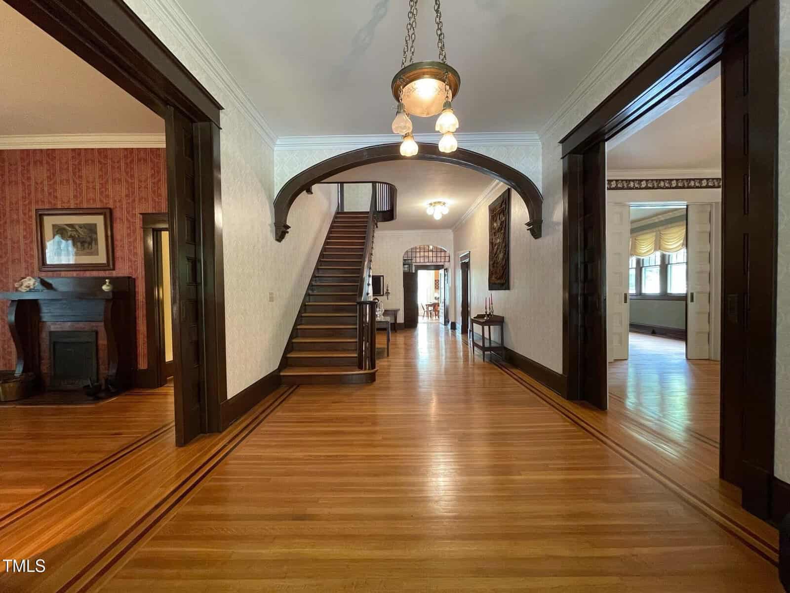 A spacious, elegant hallway with polished wooden floors and dark wood trim, featuring a staircase leading upstairs. There is a chandelier hanging from the ceiling. Doorways open into rooms on either side, with a fireplace visible to the left.