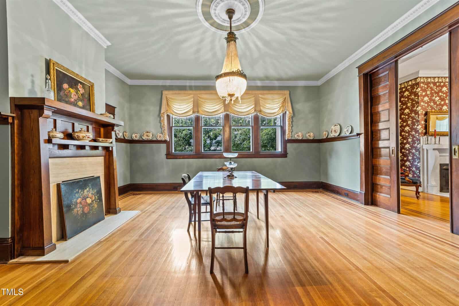 A dining room with a wooden floor featuring a table and two chairs. There's a vintage chandelier, a fireplace with art on the mantel, built-in shelves with decorative plates, large windows with curtains, and an open doorway leading to another room.