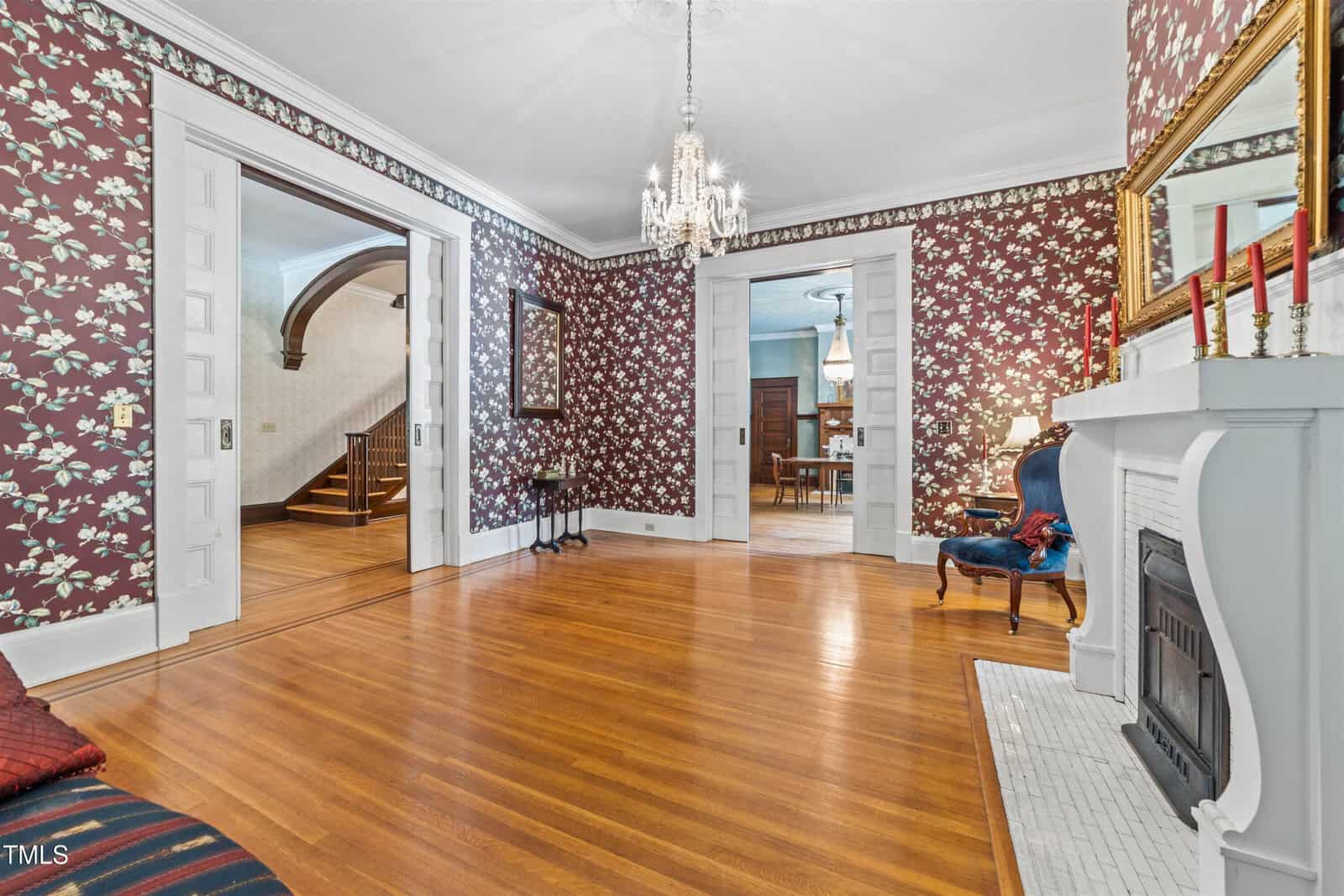 A vintage-style living room with floral wallpaper, hardwood floors, and a white fireplace. A chandelier hangs from the ceiling, and a plush blue chair sits to the right. Open doorways lead to an adjacent room and a staircase.