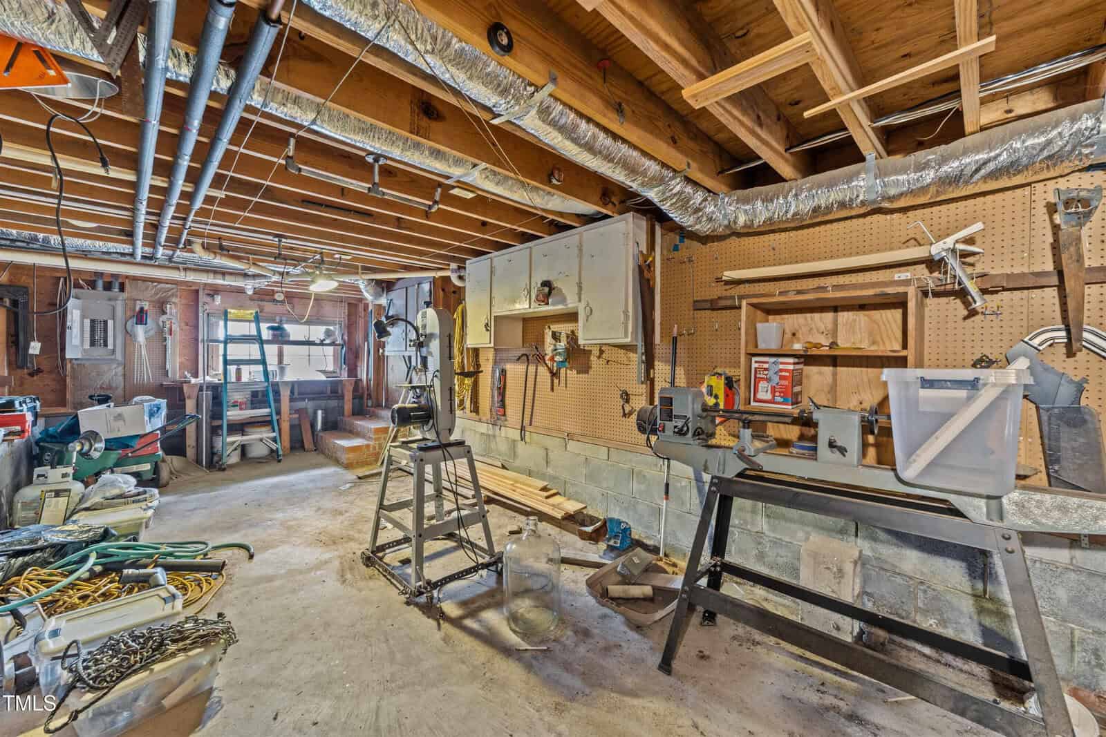 A cluttered basement workshop with exposed beams and ducts. Tools and materials are scattered on workbenches and the floor. A pegboard is mounted on the wall with various tools hanging. A step ladder and a workbench are prominently visible.