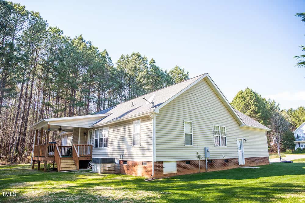 A single-story house with beige siding and a gray roof stands next to a wooded area. It has a small wooden deck at the back and a brick foundation. The lawn is green, and the sky is clear and sunny.