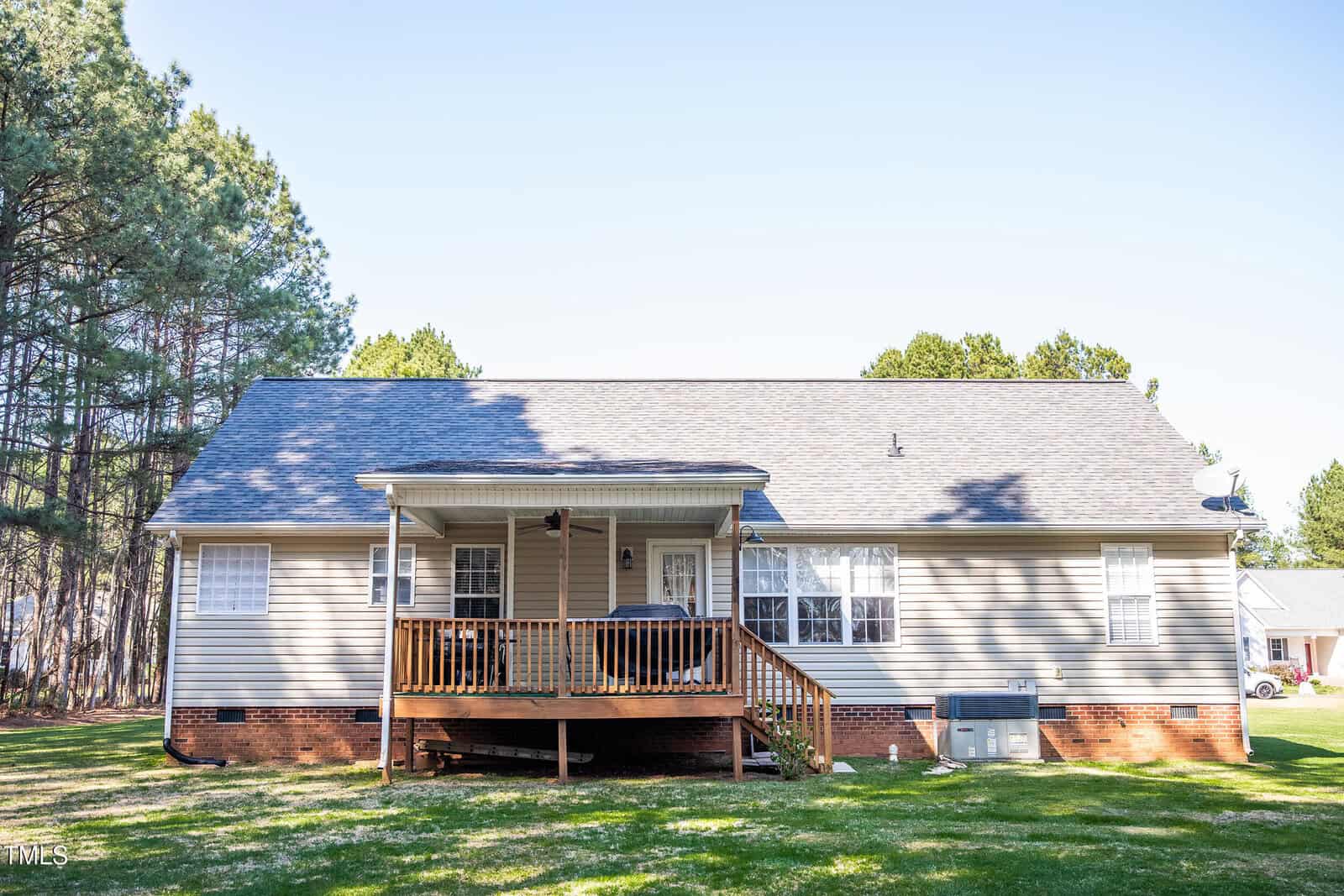A single-story house with beige siding and a dark gray roof. A wooden deck with steps extends from the back. Trees are in the background, and there is an air conditioning unit on the right side. The sky is clear.