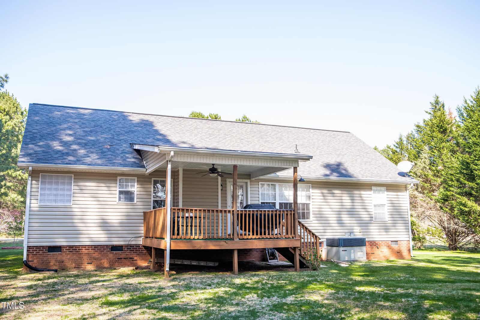 A beige house with a sloped roof and a covered wooden deck at the back. There are trees in the background and an air conditioning unit near the deck. The sky is clear and sunny.