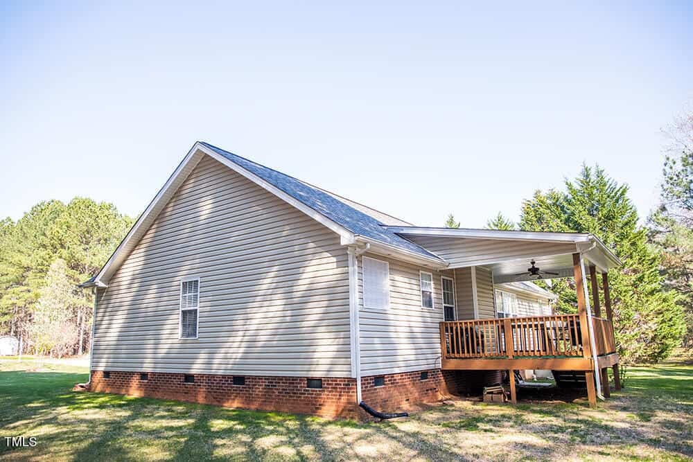 A house with light gray siding and a brick foundation features a covered wooden porch. The roof is covered in dark shingles. The surrounding area includes a grassy lawn and trees under a clear blue sky.