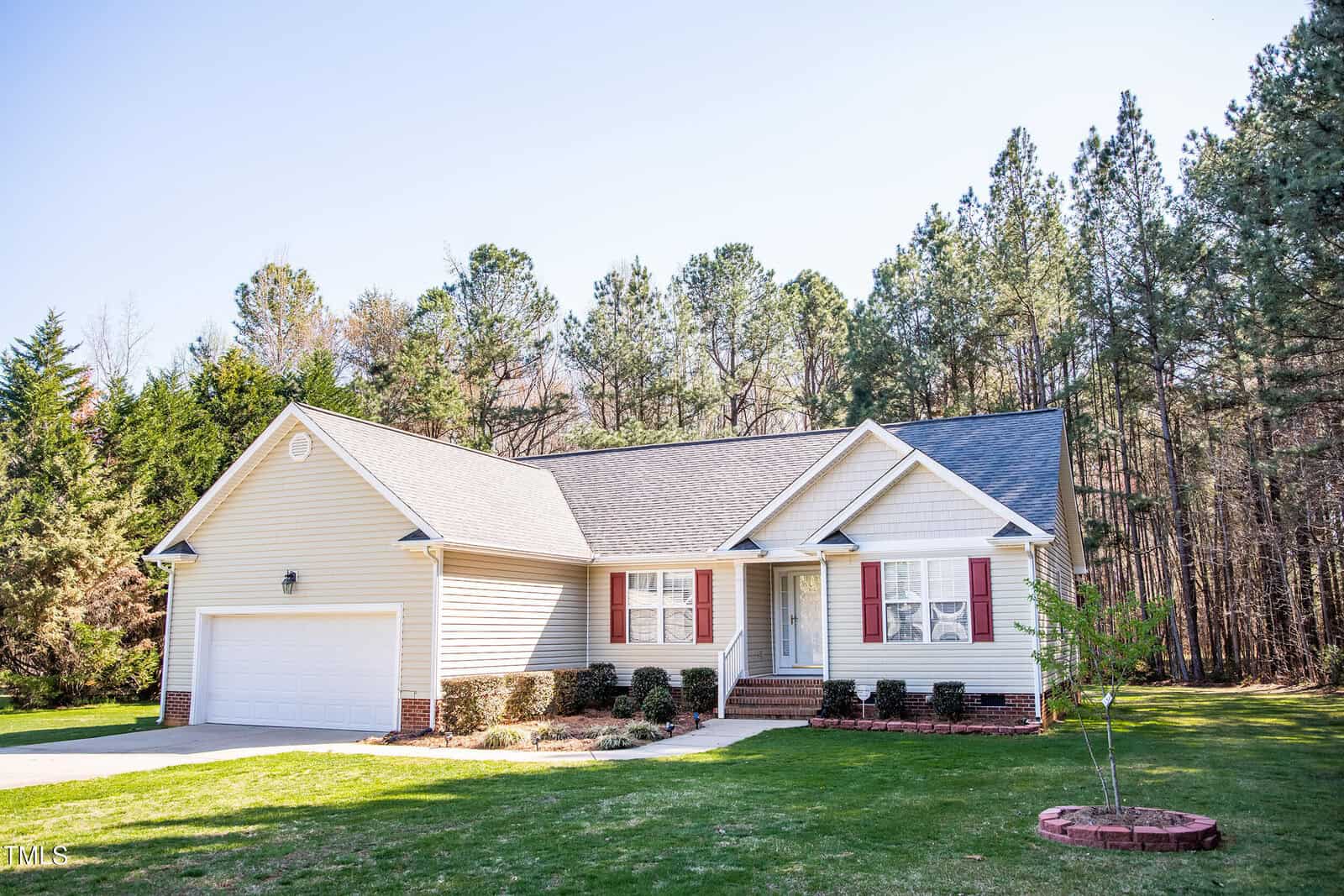 A single-story house with beige siding, red shutters, and a dark grey roof. There is a two-car garage on the left. The house is surrounded by a grassy lawn and trees in the background. A paved driveway leads to the garage.
