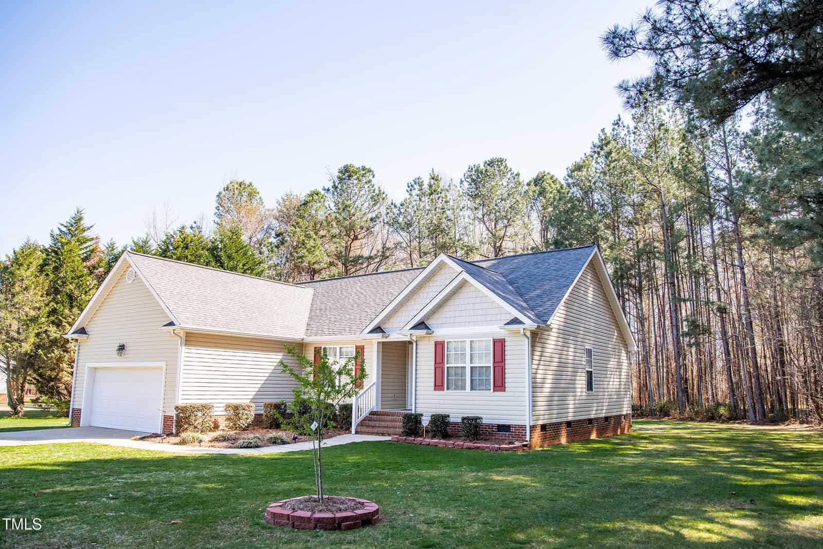 Single-story house with beige siding, red shutters, and a dark roof. It has a two-car garage and a small front porch with steps. In front, there is a small, round brick planter. The house is surrounded by trees and a lawn under a clear blue sky.