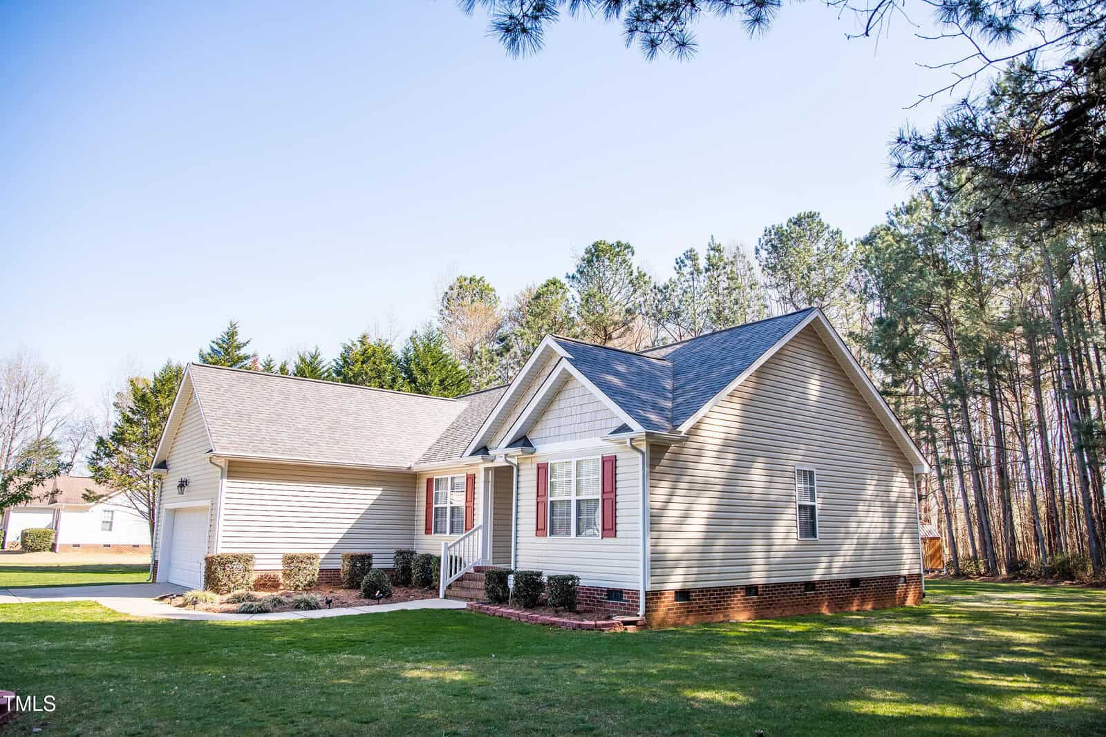 A beige, single-story house with red shutters and a gray roof is surrounded by green grass and trees. A driveway on the left leads to a two-car garage. The sky is clear and sunny.