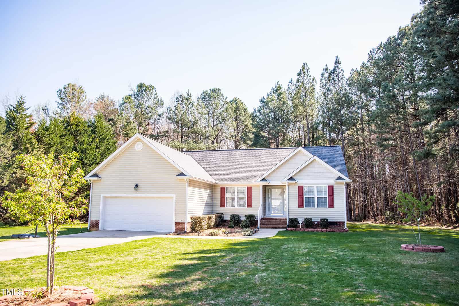 A single-story house with pale yellow siding, a gray shingled roof, and a two-car garage. It has a brick-trimmed entrance and red shutters. The house is set on a grassy lawn with trees lining the background.