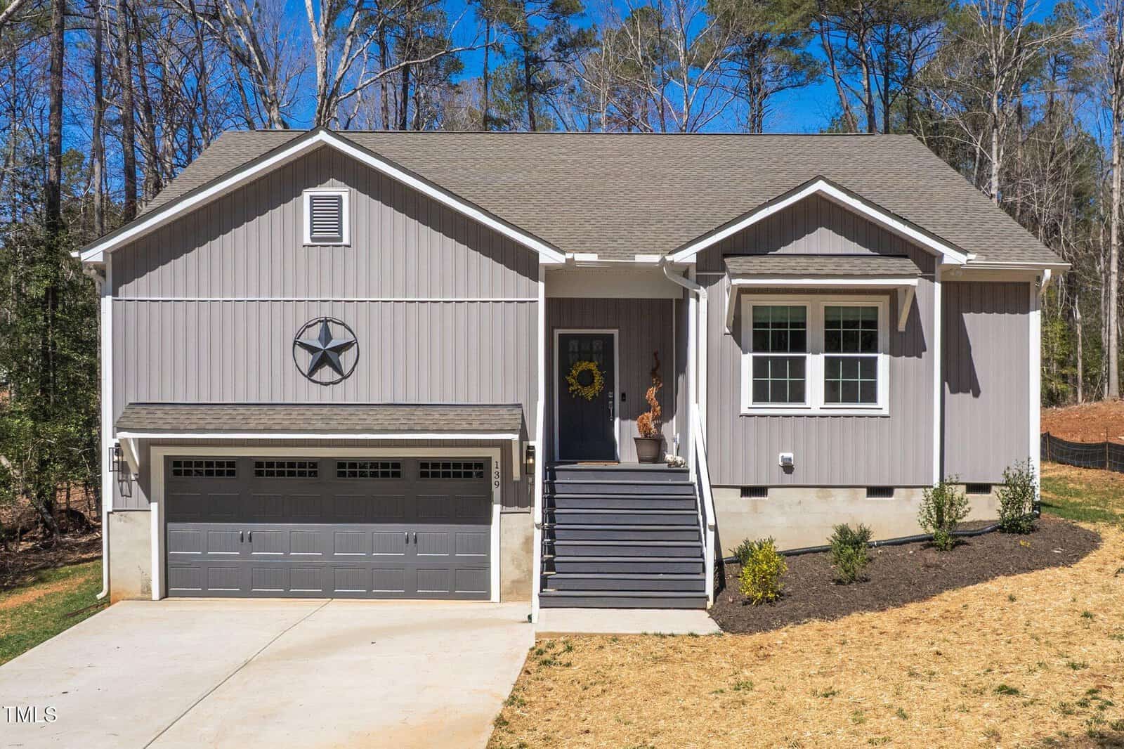 A gray single-story house with a two-car garage, front steps leading to a porch with a sunflower wreath on the door, and minimal landscaping in the front yard. Tall trees are in the background.